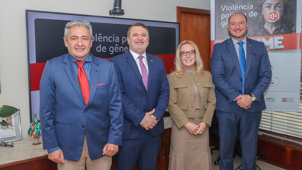 Foto de quatro pessoas em pé, uma ao lado da outra, durante o evento de lançamento na Alesc. Da esquerda para a direita, o conselheiro do TCE/SC Aderson Flores; o promotor de Justiça Fábio de Souza Trajano; a presidente do TRE-SC, Maria do Rocio; e o deputado Mauro De Nadal. Atrás deles, há banners com a identidade visual da campanha “Violência política de gênero é crime”. 