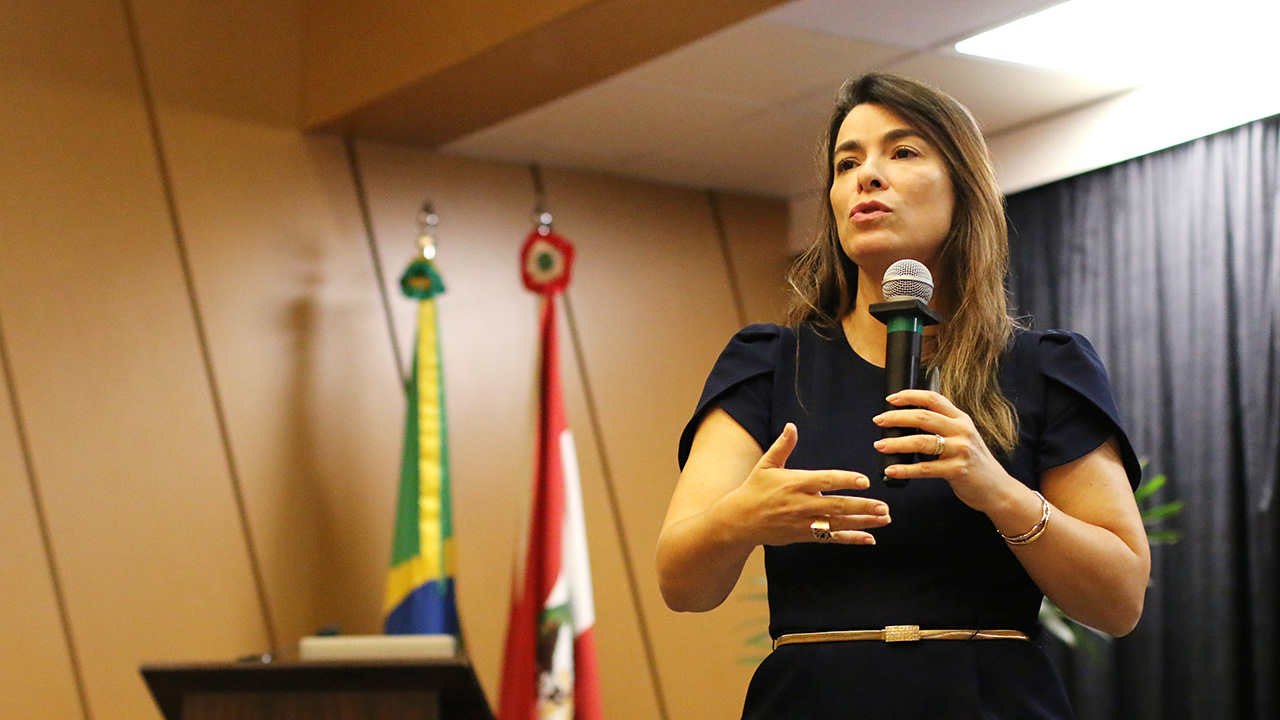 Foto da professora Cristiana Fortini, falando ao microfone, no palco do auditório do TCE/SC, durante o Congresso. Ela é uma mulher branca, de cabelos claros. Veste blusa e calça azuis. 