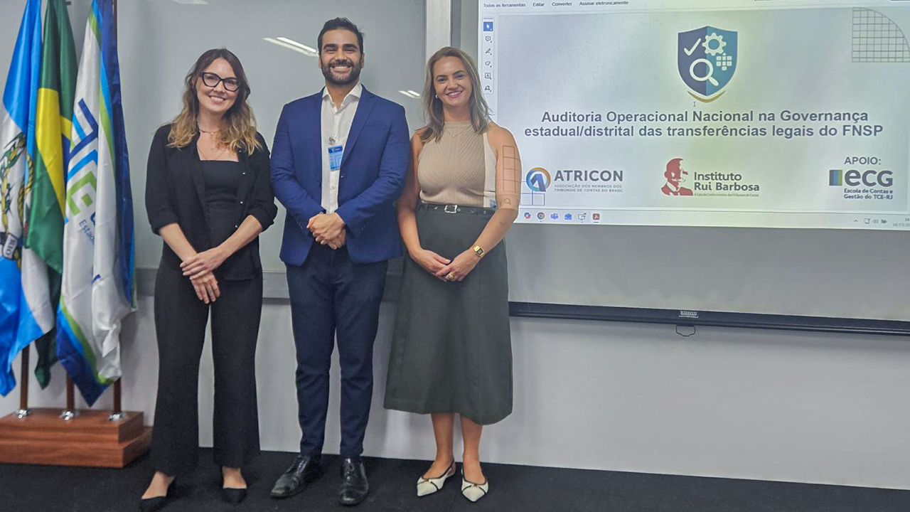 Banner horizontal com foto dos auditores fiscais de Controle Externo Rafaela Viana, Ronald Lopes e Gláucia da Cunha em pé, lado a lado, em uma sala com apresentação projetada ao fundo, no telão. Eles sorriem. A apresentação tem como título “Auditoria Operacional Nacional na Governança estadual/distrital das transferências legais do FNSP”.