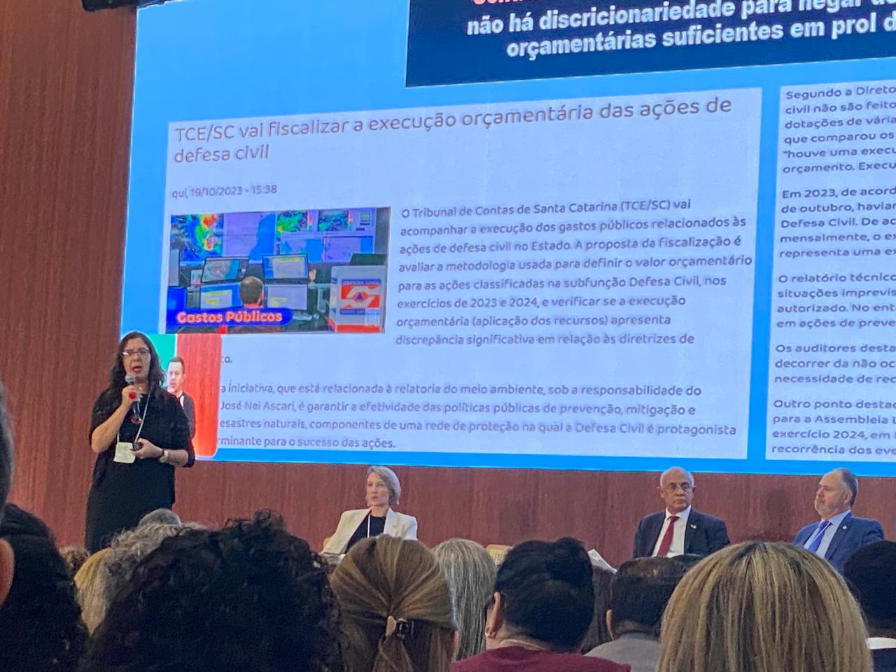 Foto da procuradora do MPC-SP Élida Graziane Pinto falando ao público durante o evento, no auditório. Ela está em pé, ao fundo, próxima ao telão que projeta a sua apresentação. À frente dela, de costas, o público que a assiste. Na projeção, o título "TCE/SC vai fiscalizar a execução orçamentária das ações de defesa civil".