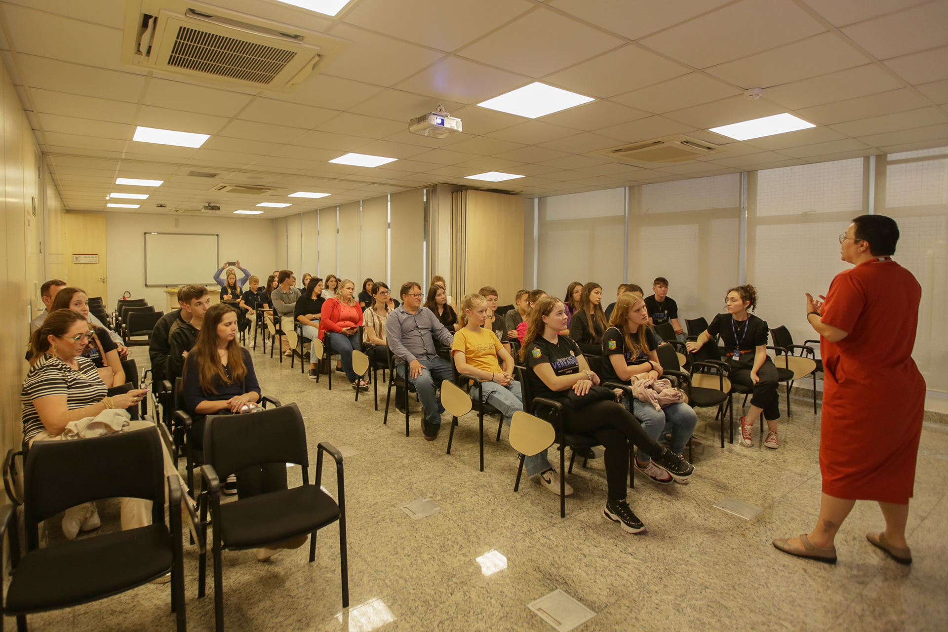 Foto dos vereadores mirins em uma sala de aula do Tribunal de Contas. Eles estão sentados em cadeiras enfileiradas. À frente deles, uma mulher faz uma explanação. 