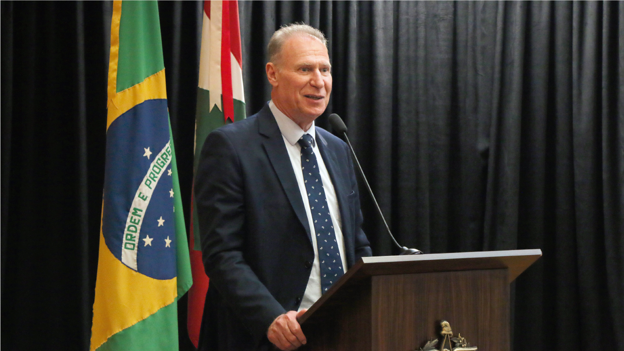 Foto do presidente do TCE/SC, conselheiro Herneus De Nadal, falando ao púlpito durante a abertura da aula magna. Ele é um homem branco, de cabelos claros. Veste terno e gravata e está em pé. Atrás dele, aparecem as bandeiras do Brasil e de Santa Catarina. 