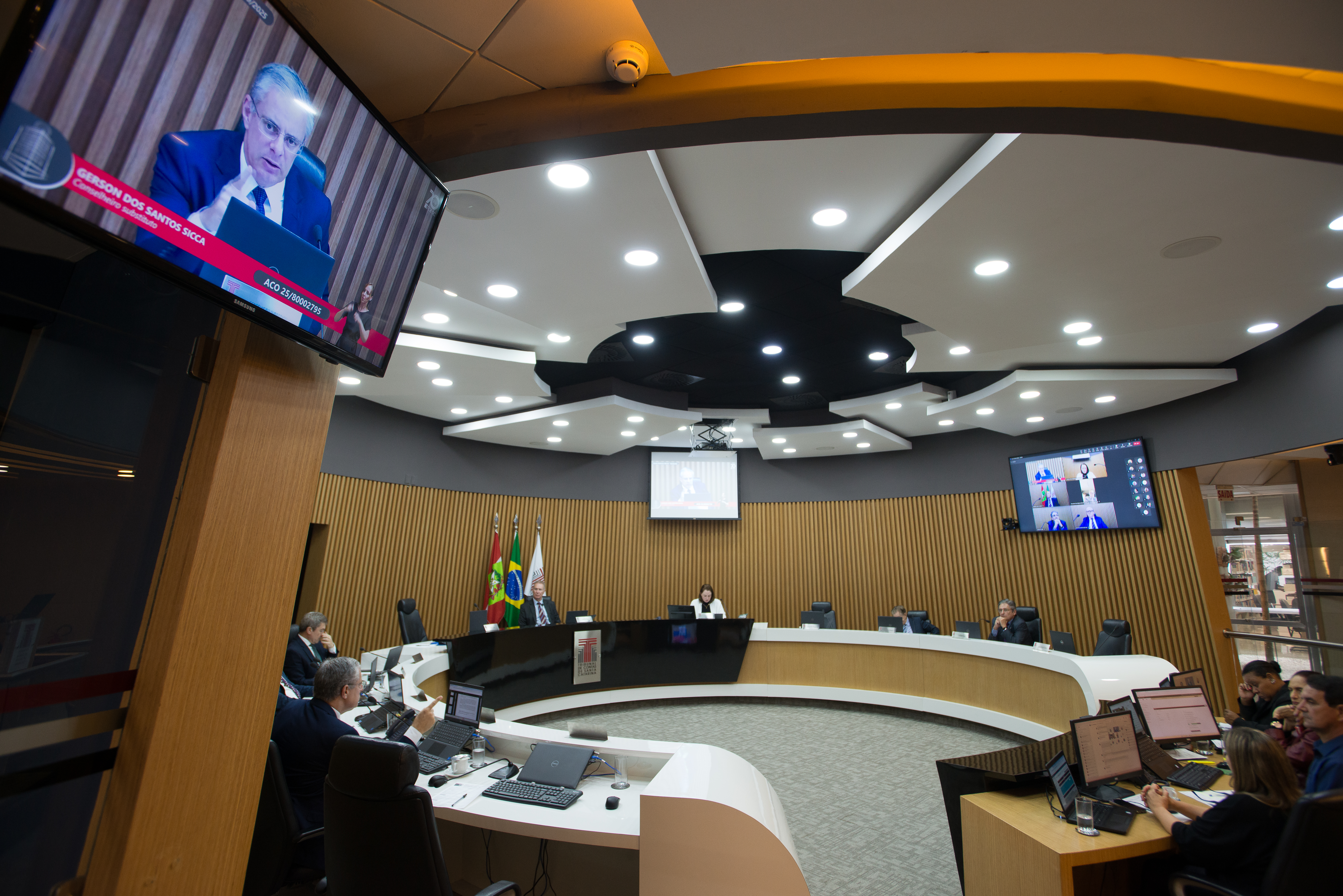 Foto horizontal do Plenário do TCE/SC. O ambiente possui formato semicircular, com bancadas equipadas com computadores e assentos destinados aos conselheiros, procuradores e servidores. Ao centro, encontra-se a mesa principal da sessão, tendo ao fundo as bandeiras do Brasil, de Santa Catarina e do TCE/SC. No canto superior esquerdo, um monitor exibe a participação remota do conselheiro substituto Gerson Sicca. 