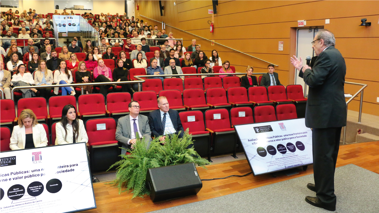 Em primeiro plano, de costas para a foto, o conselheiro aposentado Sebastião Helvécio proferindo a aula magna. Ele está em pé, no palco do auditório do TCE/SC. Ao fundo, em segundo plano e de frente, a plateia que lota o auditório e assiste à conferência. Entre o público, os conselheiros Adircélio de Moraes e Herneus De Nadal, sentados à frente do palco.  