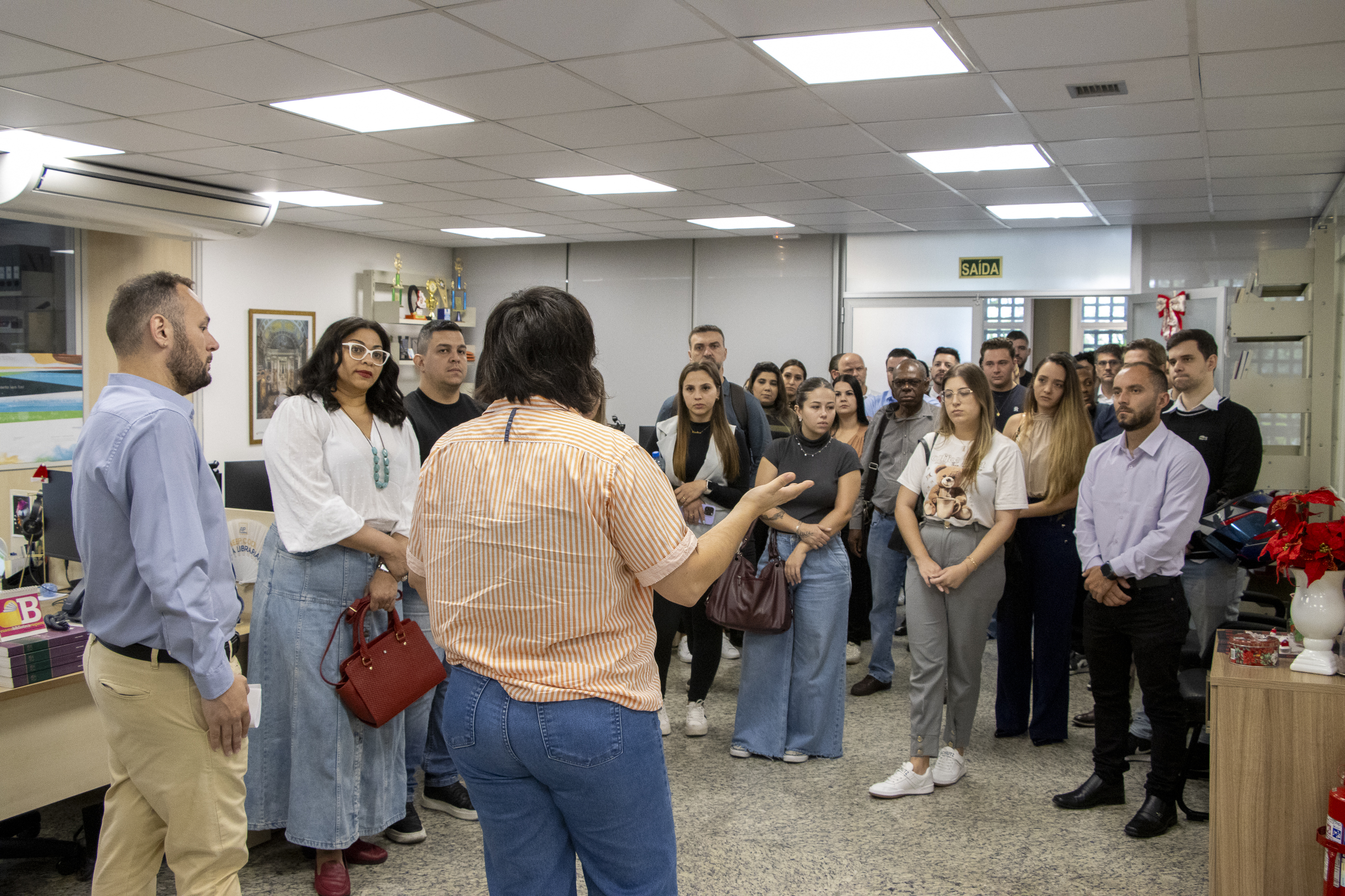 Imagem horizontal que mostra um grupo de estudantes e adultos reunidos em uma sala do Tribunal de Contas de Santa Catarina durante visita institucional. No centro, uma pessoa está de costas para a câmera enquanto explica algo ao grupo, que escuta atentamente ao redor.