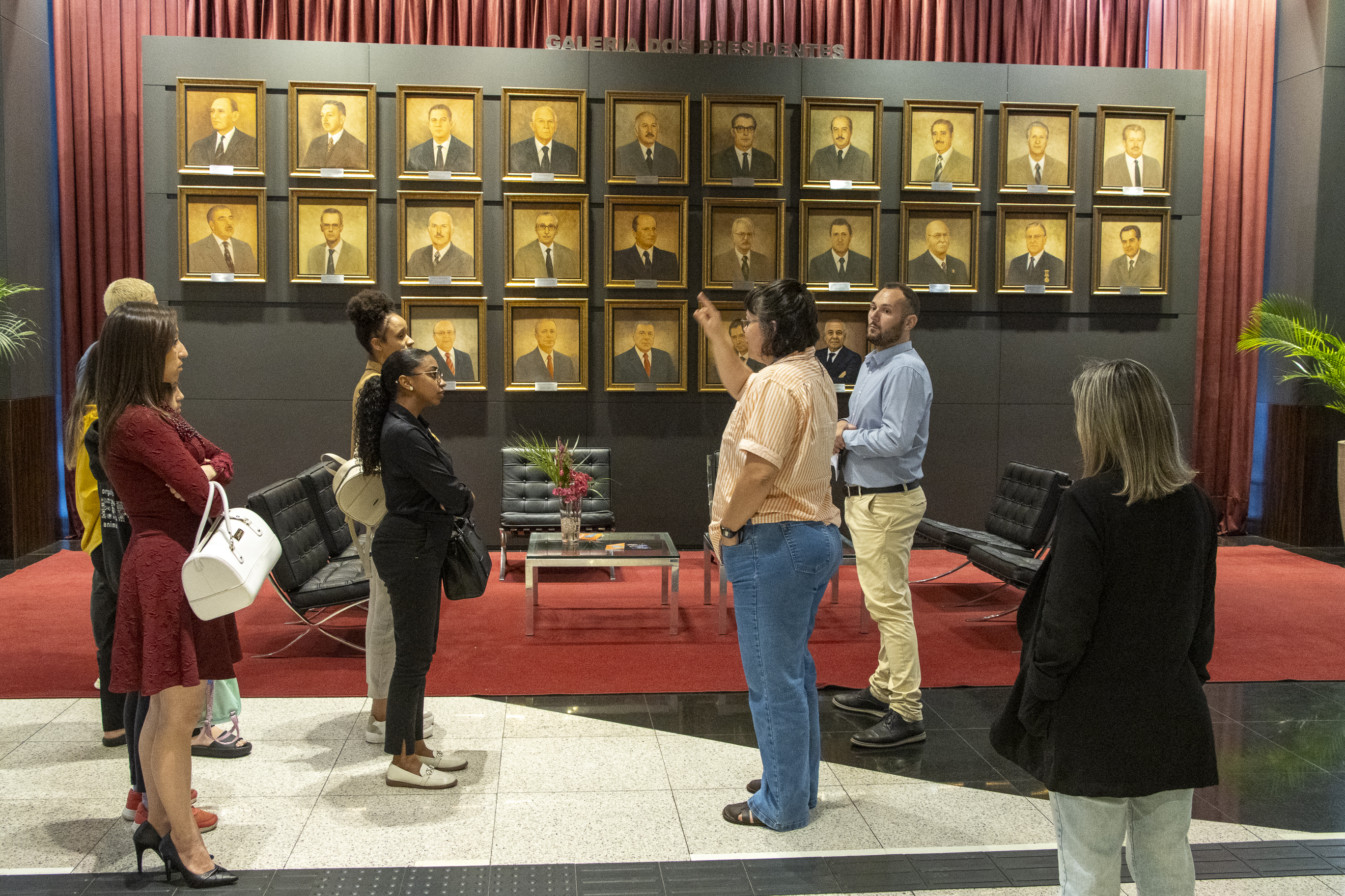 Imagem horizontal com foto de um grupo de estudantes e adultos em visita ao Tribunal de Contas de Santa Catarina, reunidos em frente à galeria de presidentes da Instituição. Uma pessoa conduz a explicação enquanto aponta para os quadros com retratos de ex-presidentes do Tribunal dispostos na parede ao fundo. O grupo observa atentamente.