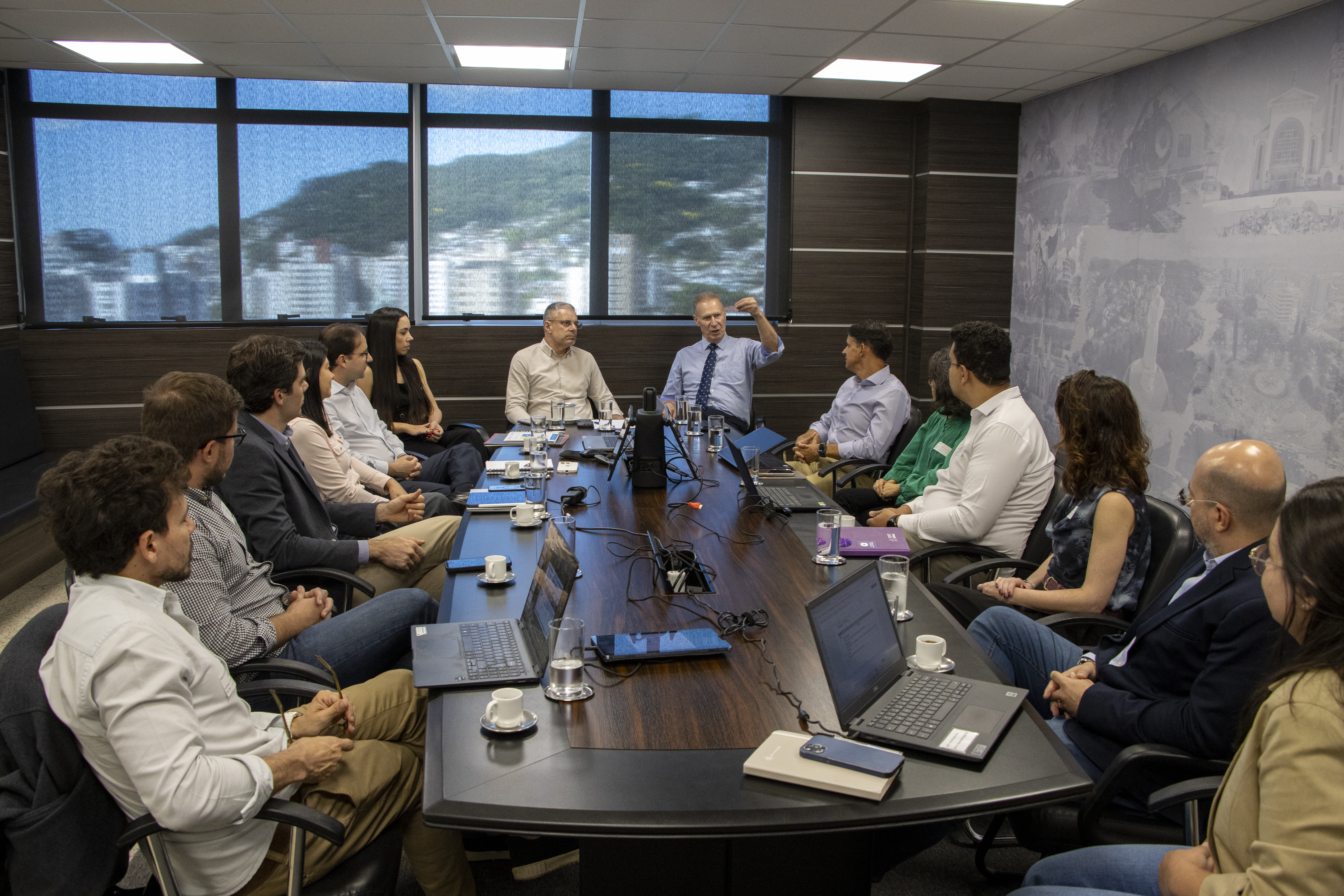Imagem mostra os participantes da reunião do Comitê Estratégico do Projeto de Apoio do Controle Externo à Gestão da Educação, que ocorreu em novembro de 2025. Há 14 pessoas sentadas ao redor de uma mesa marrom, onde há computares, xícaras, copos e celulares. Ao centro, o presidente Herneus De Nadal e o conselheiro substituto Gerson Sicca. 