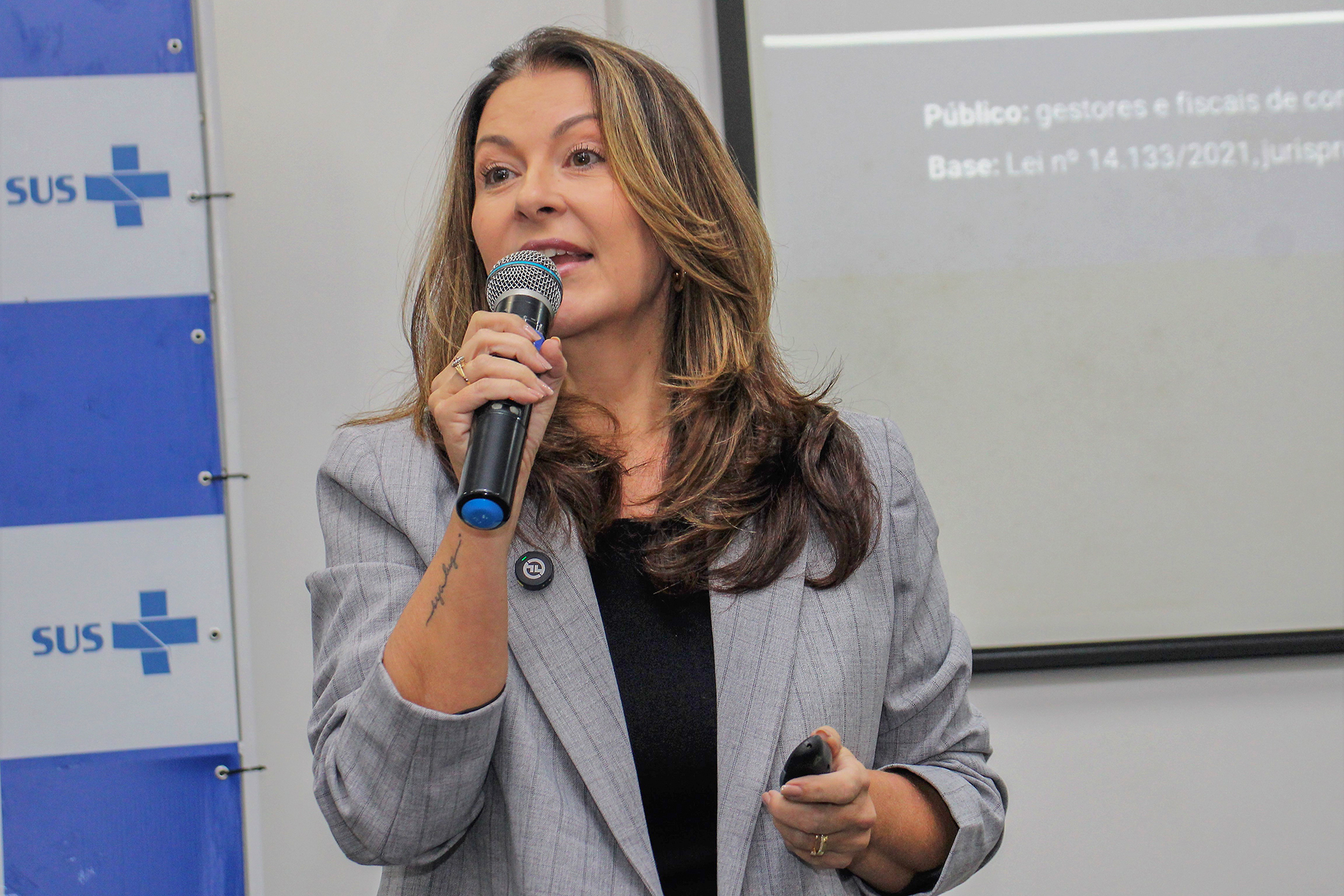 Foto da auditora Caroline de Souza, do TCE/SC, falando ao microfone durante palestra. É uma mulher branca, de cabelos castanhos. Está em pé, em frente a telão com projeção de slides.