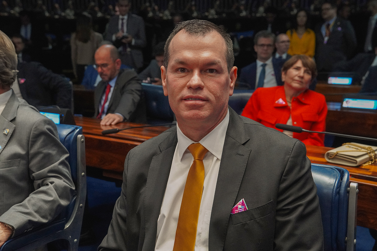 Foto do conselheiro Adircélio de Moraes Ferreira Júnior acompanhando a sessão. Ele é um homem branco, de cabelos escuros. Veste terno e gravata e está sentado ao lado das demais autoridades no Plenário do Senado. Sorri para a foto.