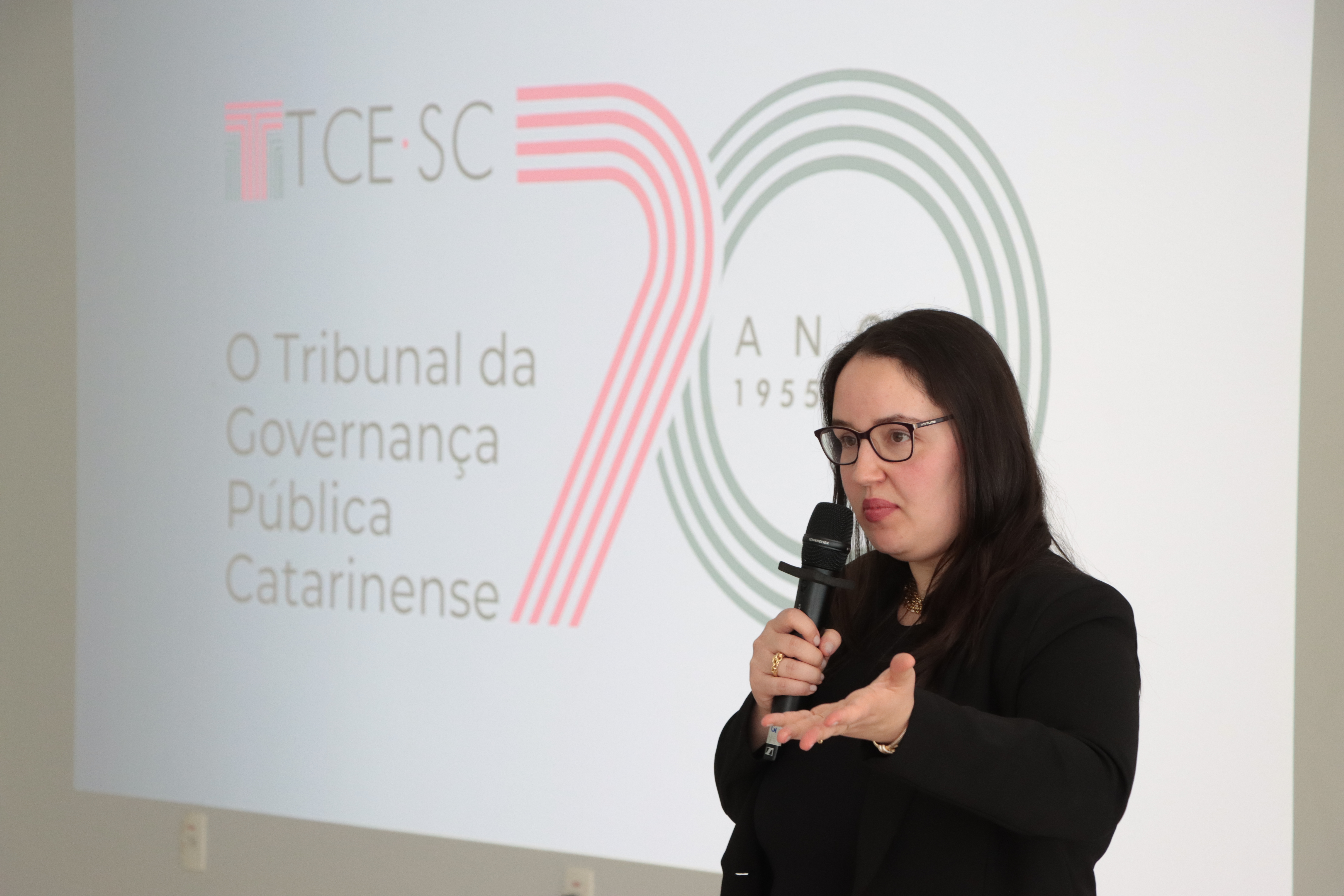 Foto da coordenadora de obras e serviços de engenharia da DLC Renata Ligocki Pedro. Ela está palestrando em frente ao banner com a logomarca do TCE.