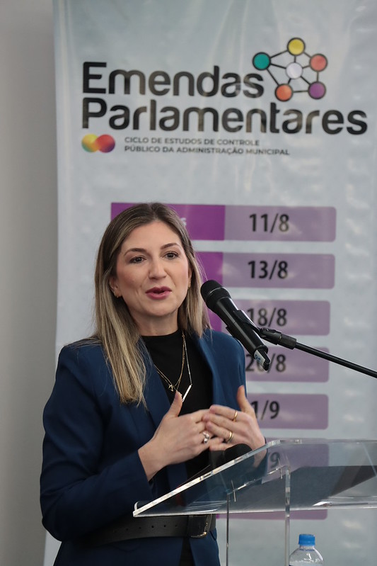 Foto horizontal da diretora do Icon, Marina de Miranda. Ela é uma mulher branca, de cabelos claros. Veste roupa azul e fala ao microfone no púlpito. Ao fundo, banner com logo do ciclo.