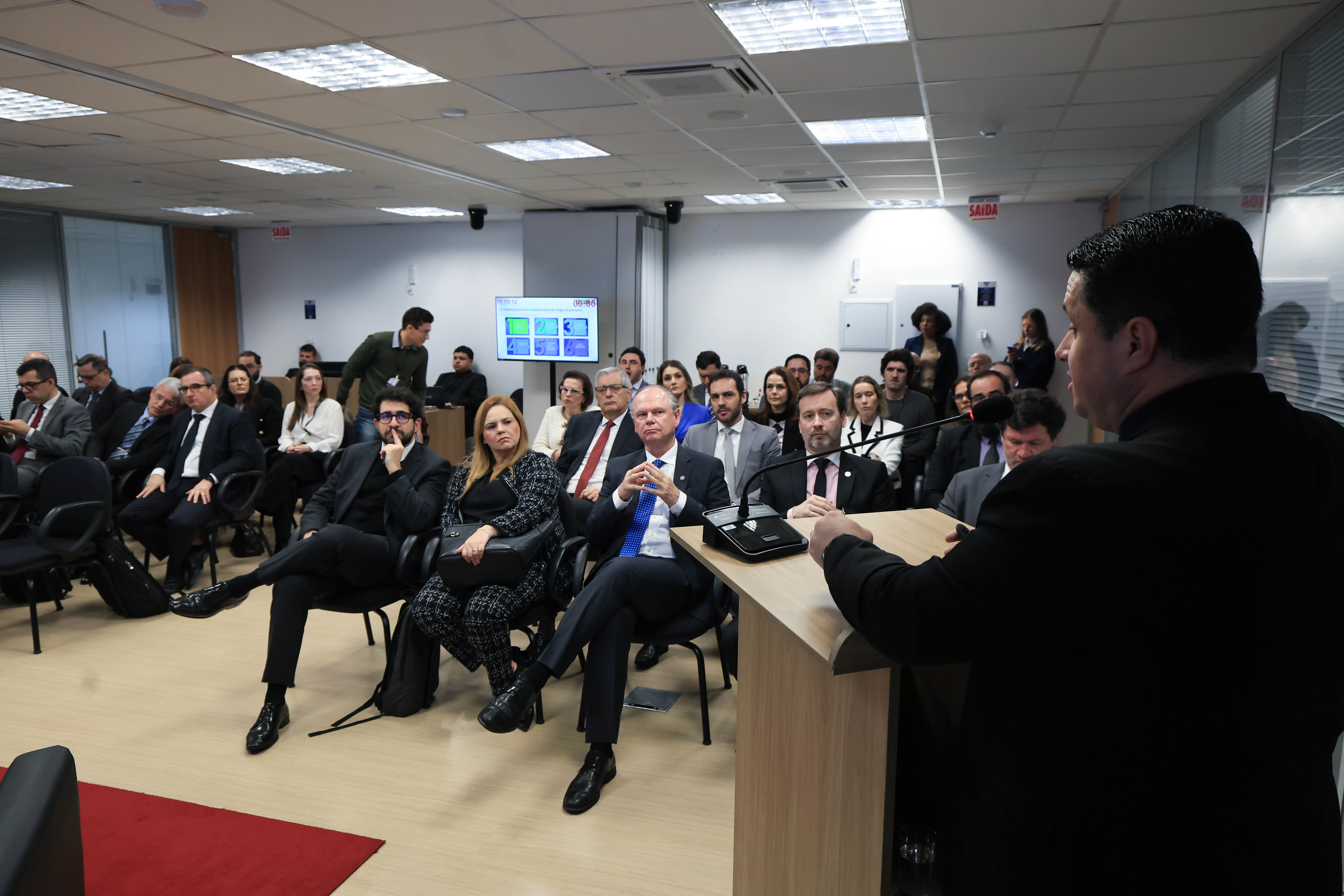 Foto horizontal do assessor Flávio Alves, em primeiro plano, de costas, falando ao microfone. Ao fundo, de frente para a foto, a plateia que o assiste, com pessoas sentadas lado a lado em cadeiras, acompanhando atentamente a fala.