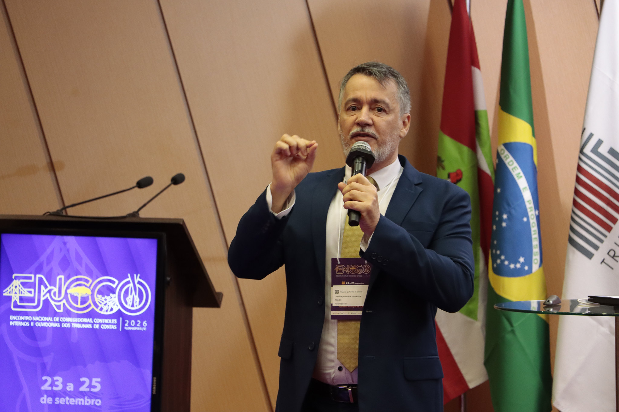 Foto horizontal do servidor Rogério Guilherme de Oliveira falando ao microfone durante o evento. É um homem branco, de cabelos e barba grisalhos. Veste terno e gravata e usa crachá no pescoço. Ele está em pé, ao lado do púlpito que projeta a logo do evento. Atrás, algumas bandeiras compõem o cenário.