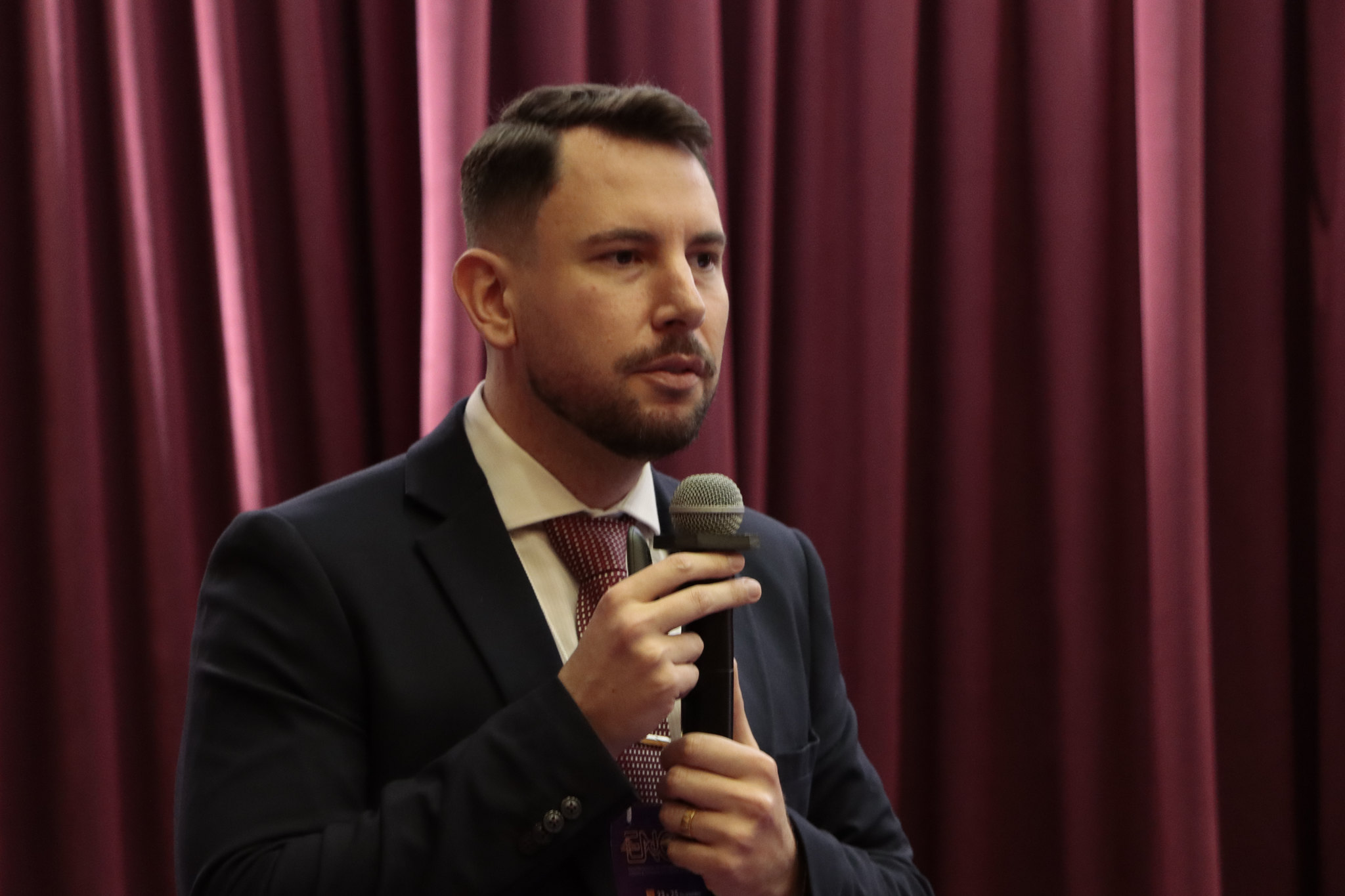 Foto horizontal do servidor Geovane Eziel falando ao microfone durante o evento. Ele é um homem branco, de cabelos e barba escuros. Veste terno e gravata e está em pé. Ao fundo, cortinas bordôs compõem o cenário.