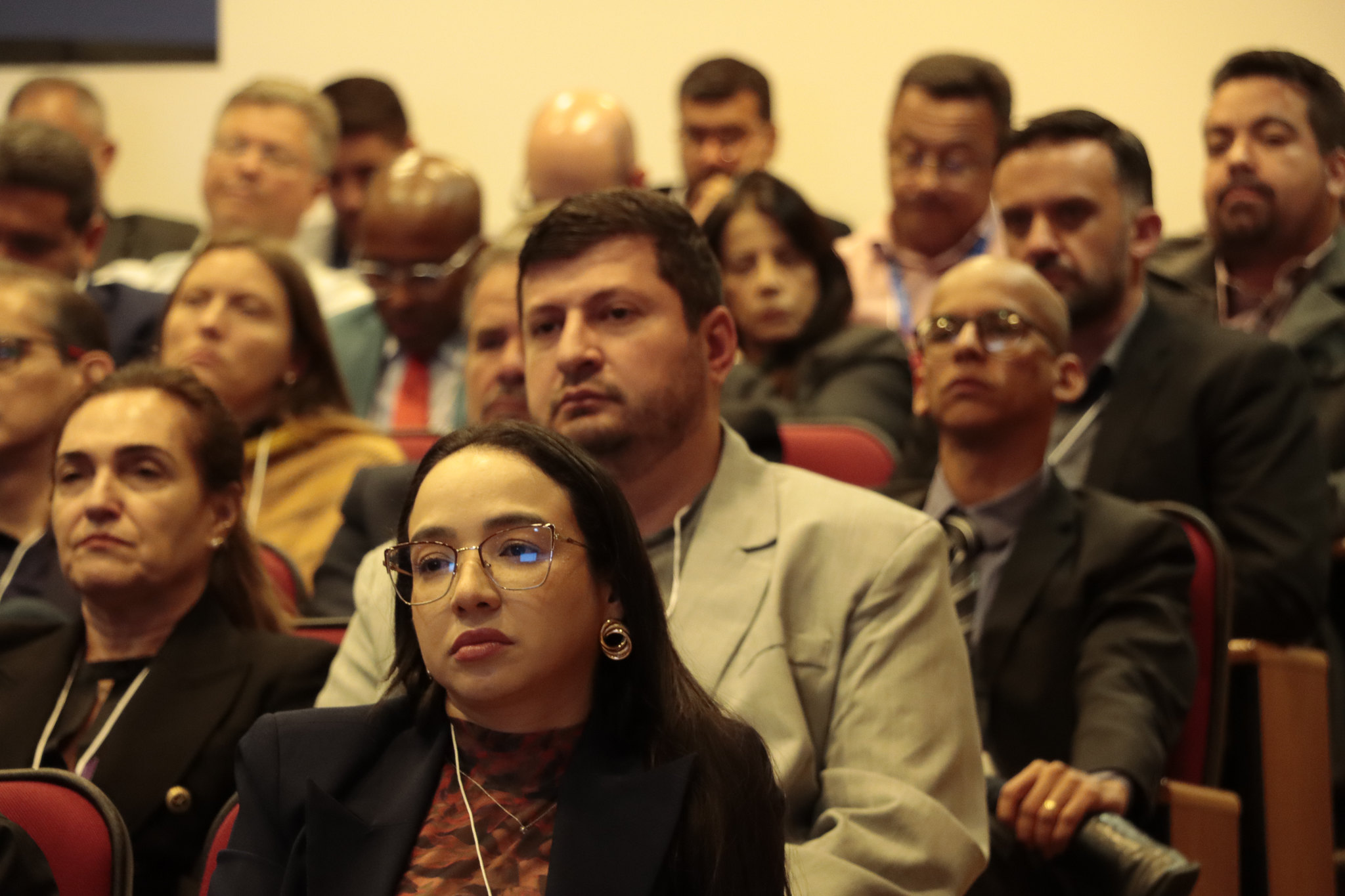 Foto horizontal de parte da plateia participante do evento. Há várias pessoas, entre homens e mulheres, sentadas em poltronas lado a lado que assistem atentamente a uma das falas.