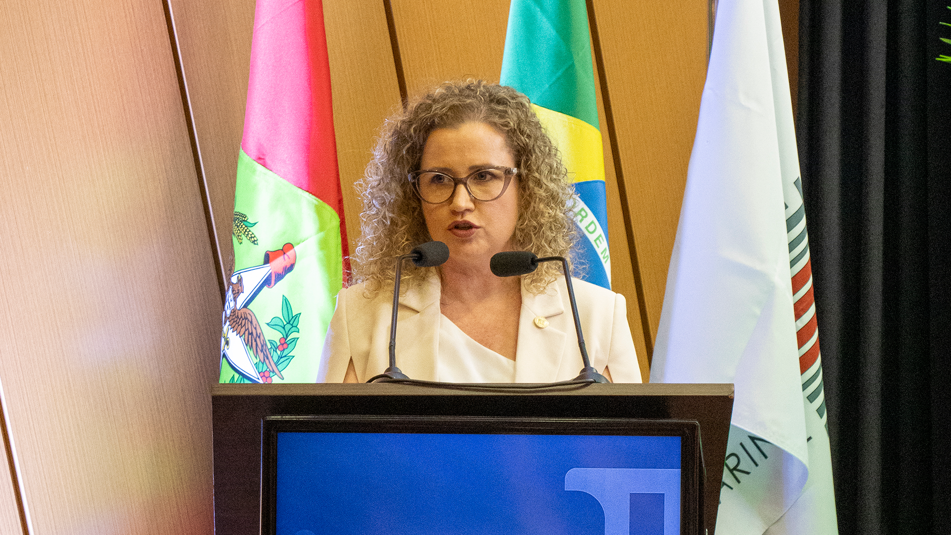 Foto horizontal da procuradora-geral do TCE/SC, Gláucia Mattjie, falando ao púlpito, durante a abertura do Enapac. Ela é uma mulher branca, de cabelos cacheados e loiros. Usa óculos de grau.