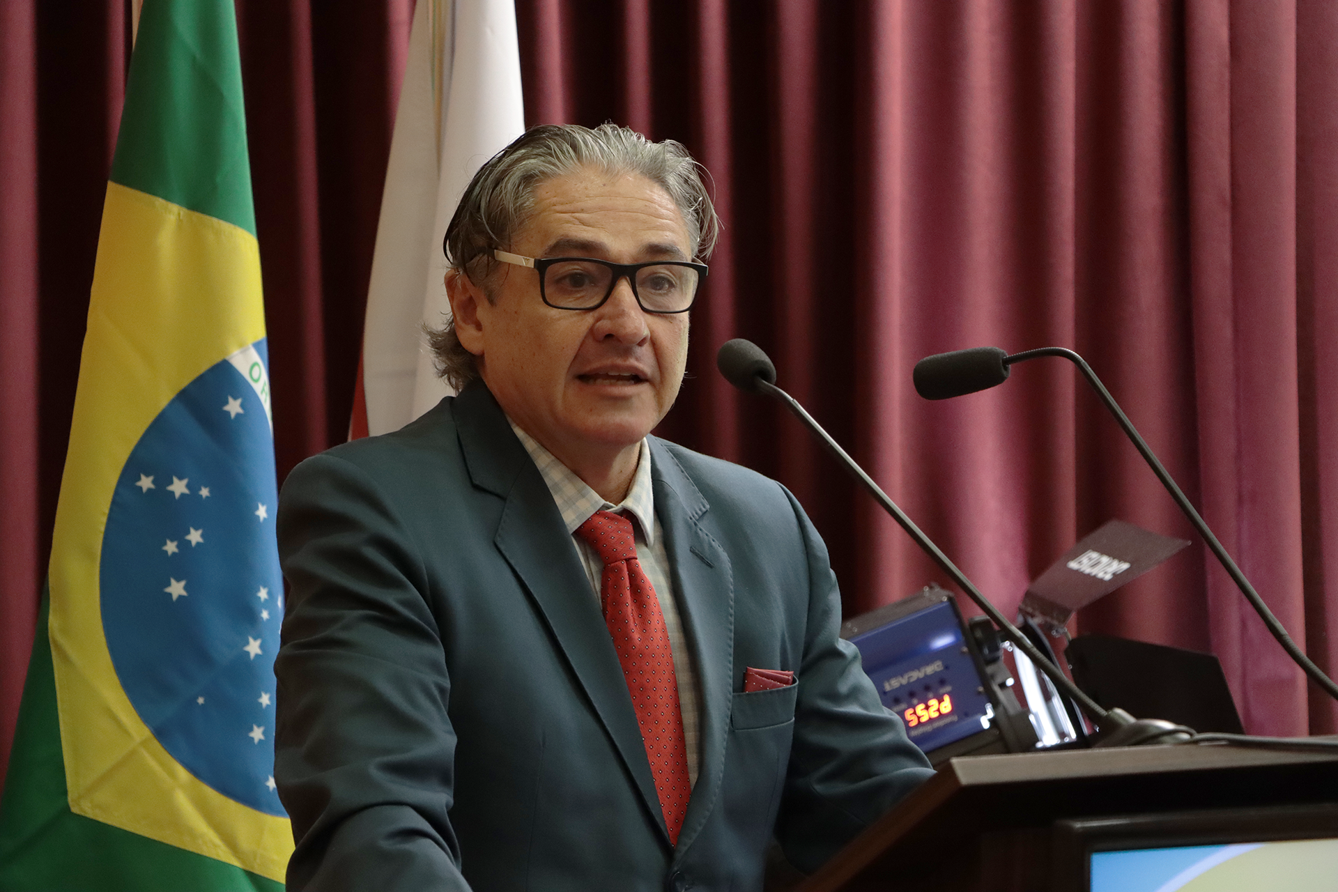 Foto do conselheiro do TCE/SC Aderson Flores falando ao microfone no púlpito, durante abertura do congresso. É um homem branco, de cabelos grisalhos. Veste terno e gravata e usa óculos de grau. Está em pé. Ao fundo, cortinas bordôs.