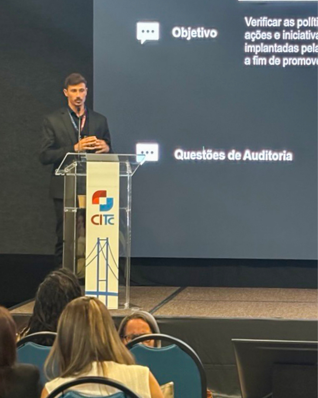 Foto do auditor fiscal de Controle Externo Alexandre Becsi durante apresentação no CITC. Ele é um homem branco, de cabelos e bigode escuros. Veste paletó preto. Está em pé, no púlpito. Ao fundo, no telão, é exibida a sua apresentação.