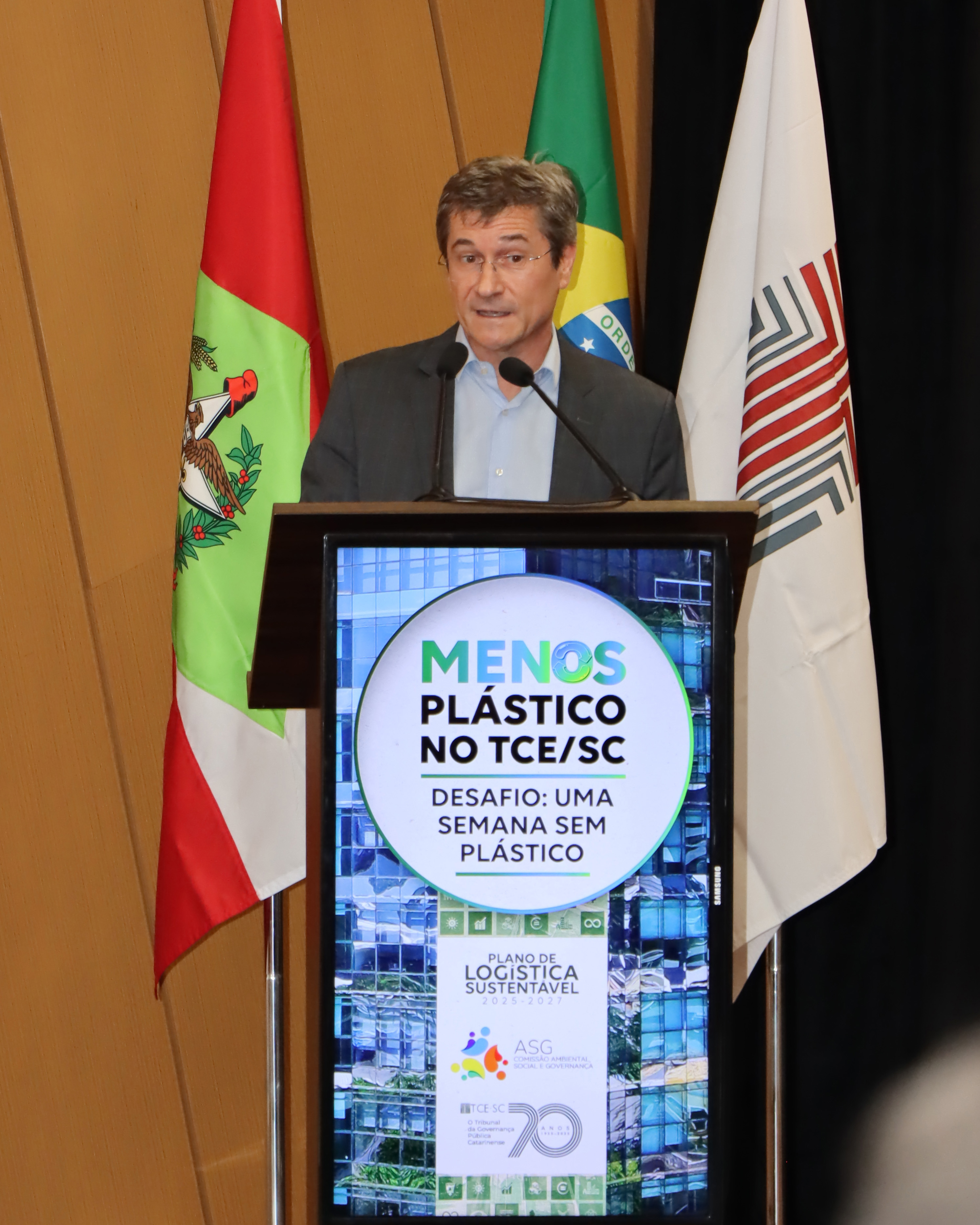 Foto do vice-presidente do TCE/SC, conselheiro José Nei Ascari, discursando no púlpito do auditório, durante o evento no Tribunal. Ele é um homem branco, de cabelos claros. Veste paletó preto e usa óculos de grau. Atrás dele, aparecem as bandeiras de Santa Catarina, do Brasil e do TCE/SC.