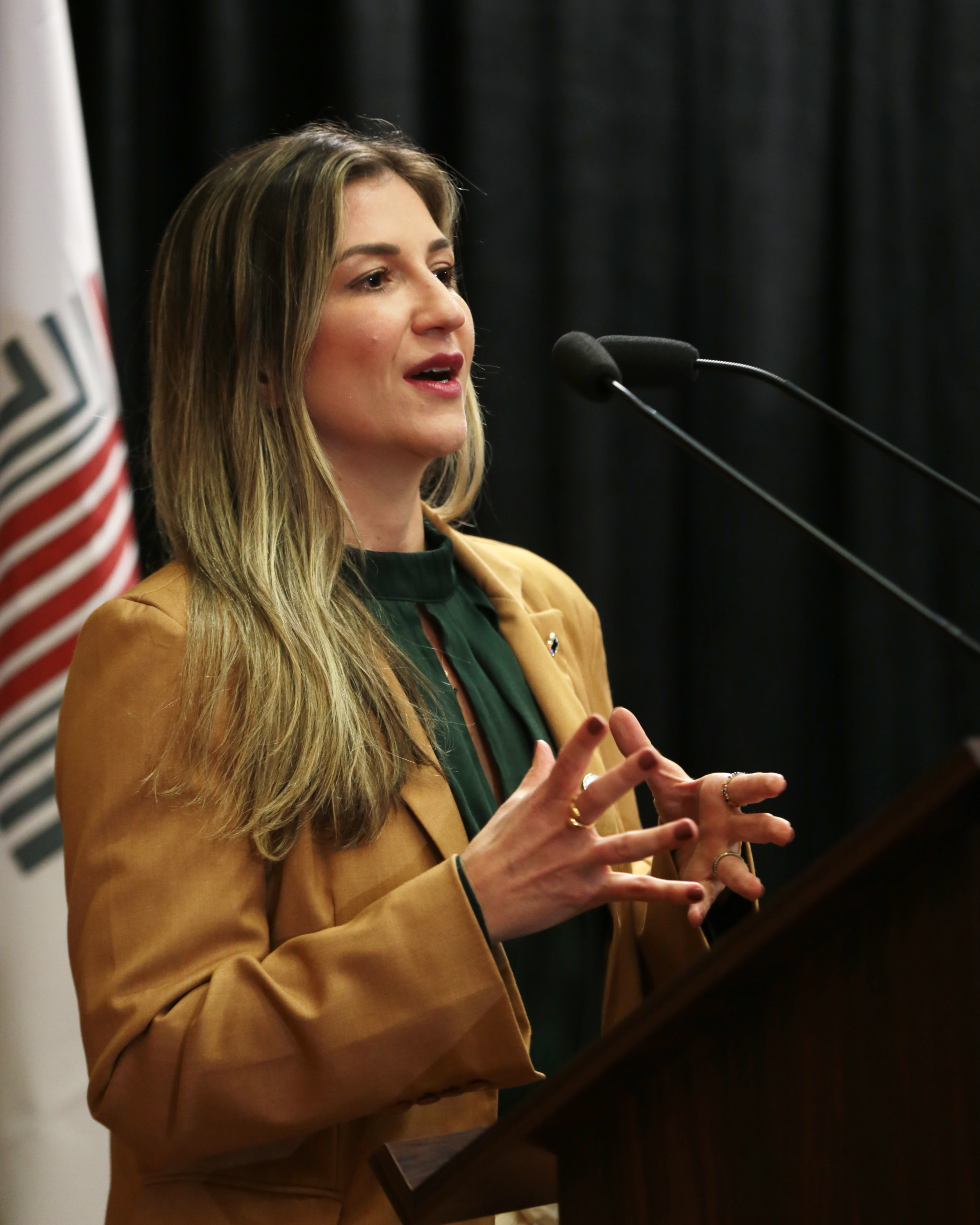 Foto da coordenadora da Comissão ASG do TCE/SC, Marina Ferraz de Miranda, discursando no púlpito, durante a abertura do evento, no palco do auditório. Ela é uma mulher branca, de cabelos loiros. Veste roupa bege e gesticula com as mãos enquanto fala.
