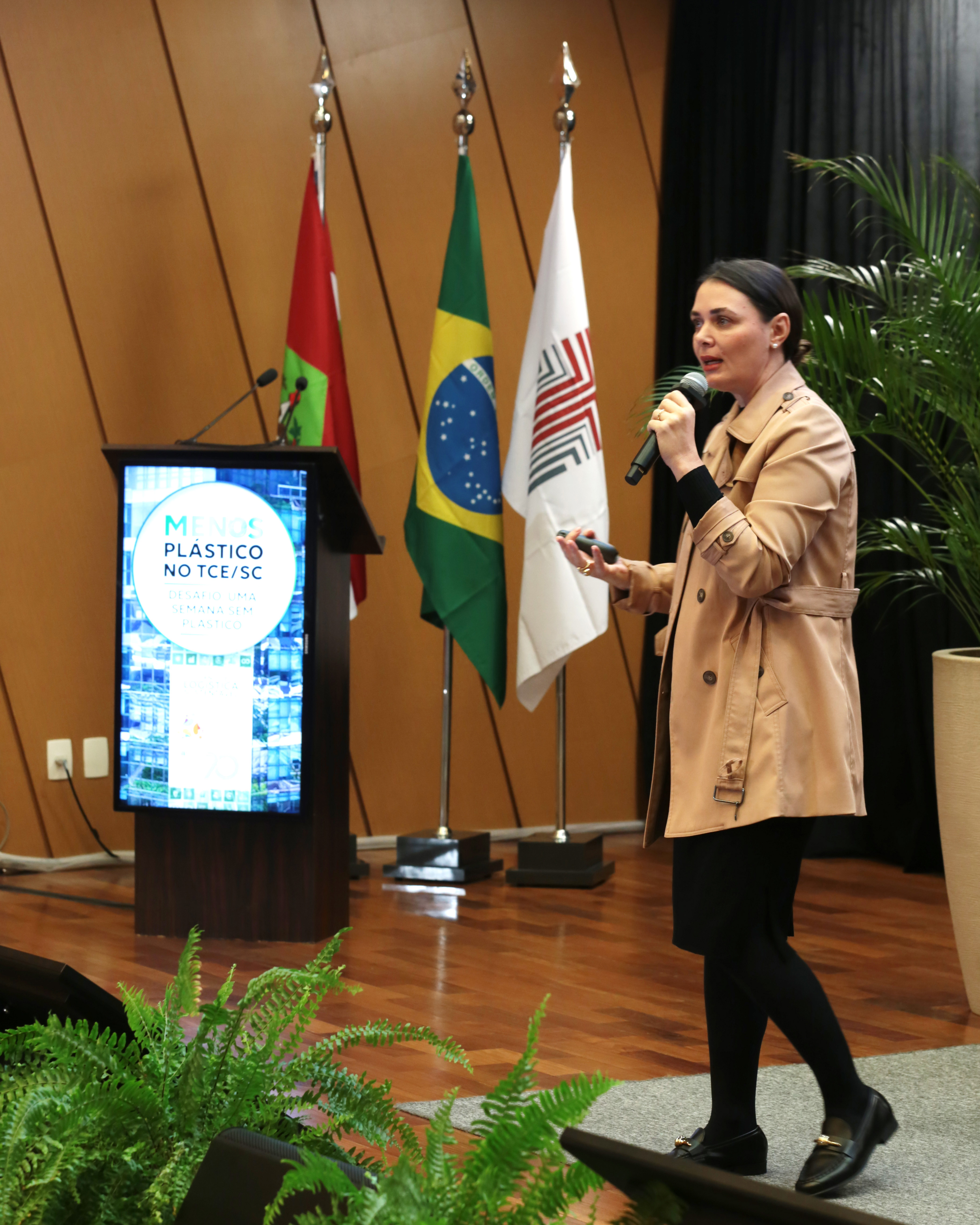 Foto da servidora no Ministério da Gestão e Inovação em Serviços Públicos Giselle Floriano Coelho palestrando durante o evento. Ela é uma mulher branca, de cabelos escuros, que estão presos. Veste um casaco bege e está em pé, no palco do auditório. Atrás dela, aparecem as bandeiras de Santa Catarina, do Brasil e do TCE/SC.