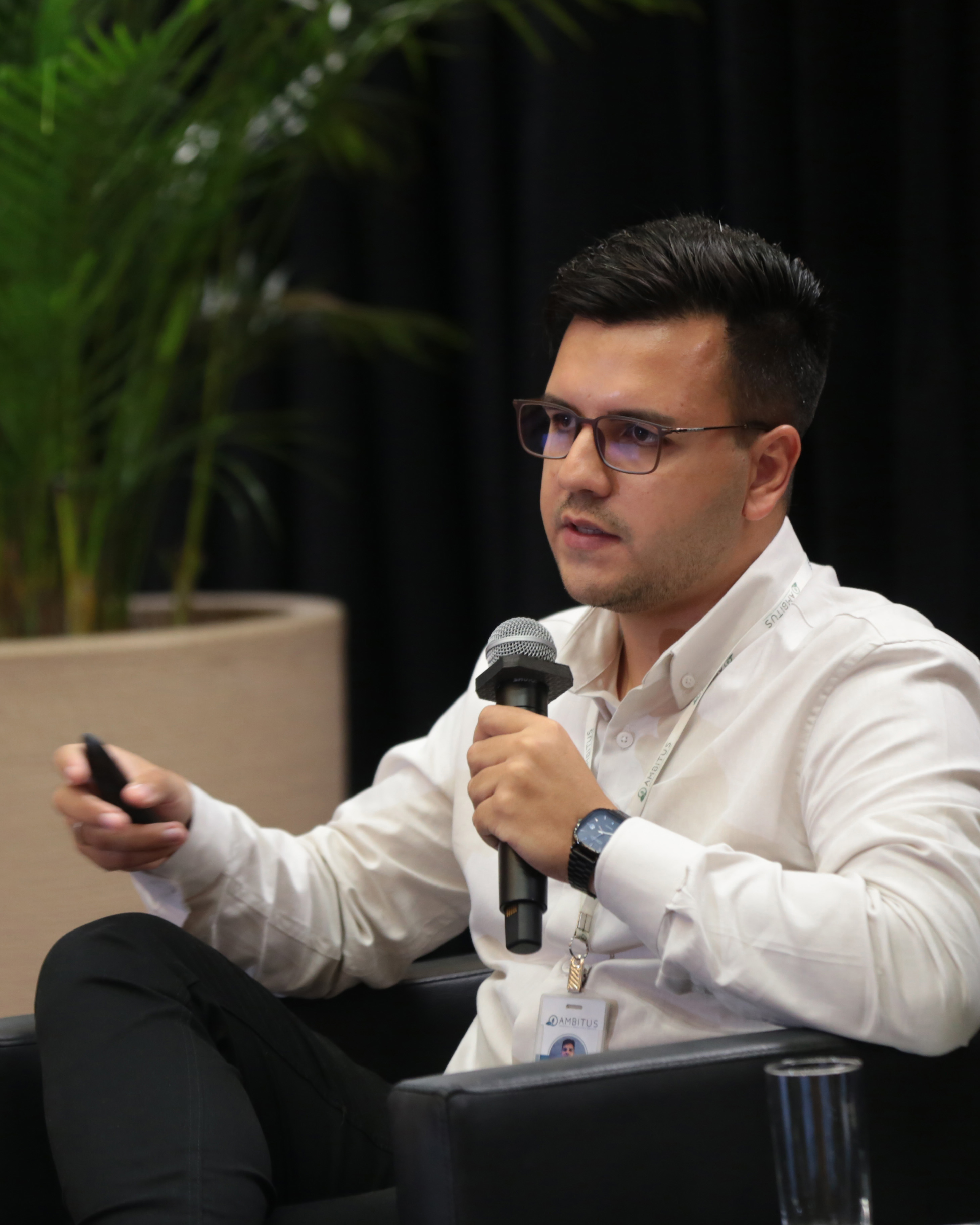 Foto do palestrante Marcos Romero Fogaça, gerente de Projetos e responsável técnico da empresa Ambitus, falando ao público, no palco do auditório. Ele é um homem branco, de cabelos escuros. Veste camisa branca, usa óculos de grau e está sentado em uma poltrona. 
