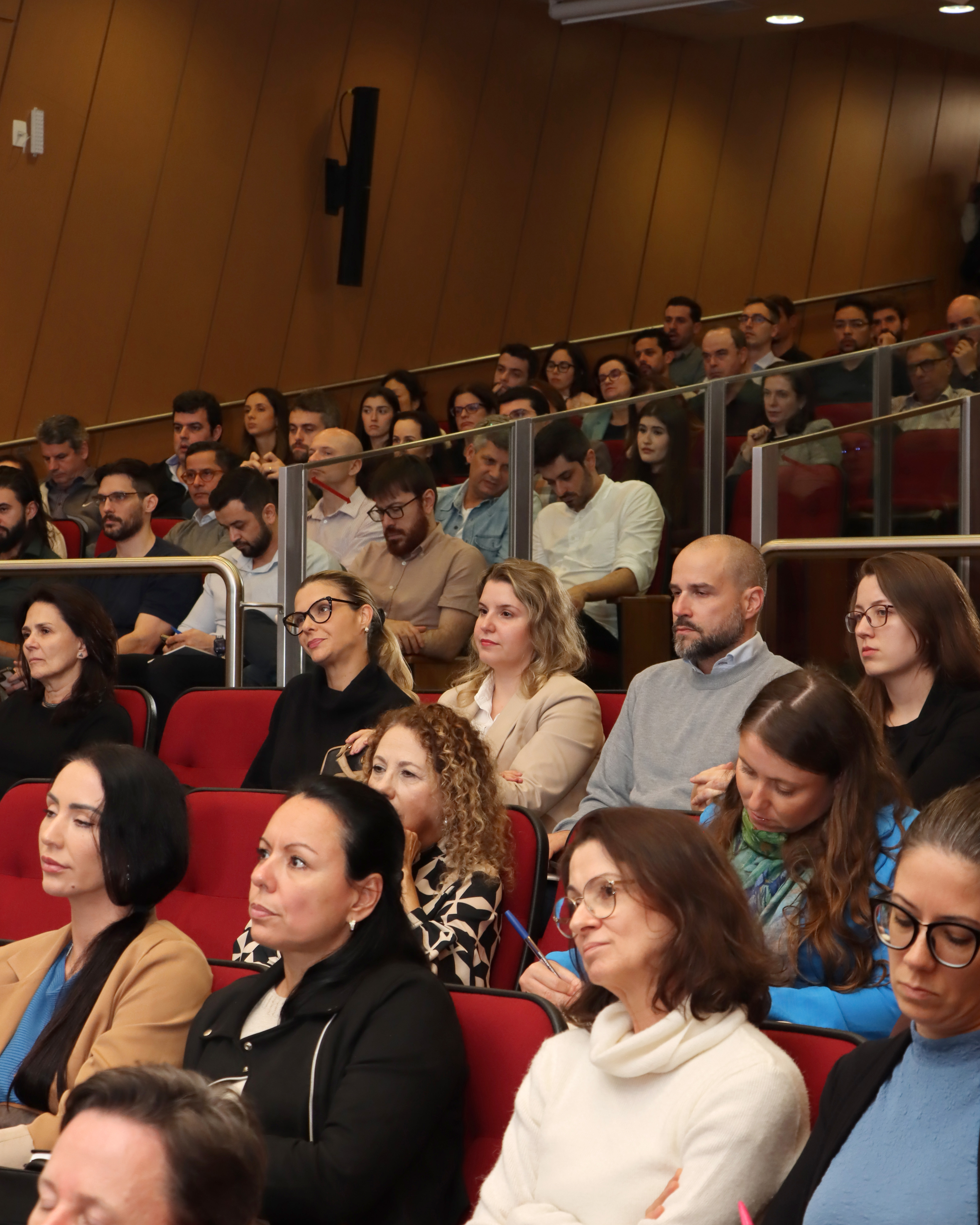 Foto de parte da plateia durante o evento. Há várias pessoas, sentadas em poltronas vermelhas do auditório, uma ao lado da outra, assistindo às palestras.