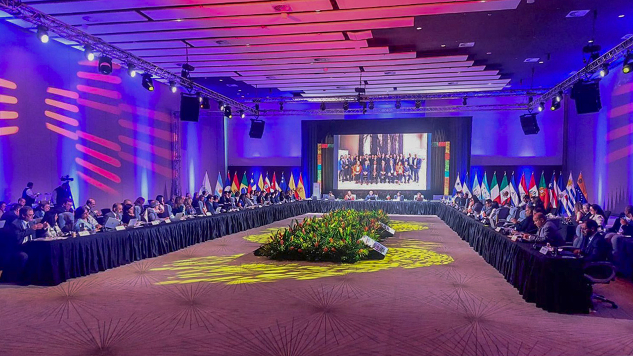 Foto do Salão principal da Assembleia da Olacefs. Uma grande mesa em formato "U", onde estão sentados diversos representantes dos TCs.
