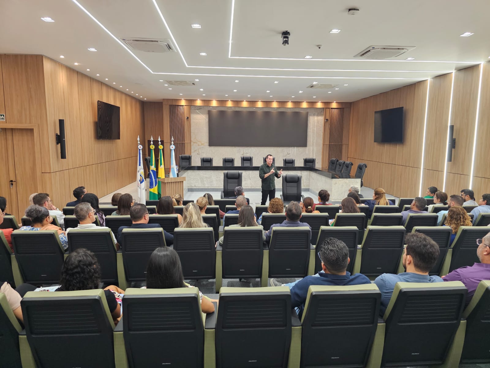 Foto horizontal que mostra o conselheiro Adircélio de Moraes Ferreira Júnior (ao fundo) falando aos servidores do TCE/AC. Ele está em pé, com o microfone nas mãos, no plenário. À frente dele, de costas para a foto, os servidores que assistem à sua fala, sentados em cadeiras, um ao lado do outro. 