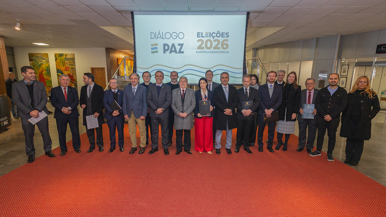 Foto horizontal de várias autoridades reunidas, lado a lado. Entre os presentes, o conselheiro Aderson Flores. Ao fundo, no telão, a inscrição "Eleições 2026, Diálogo Paz".