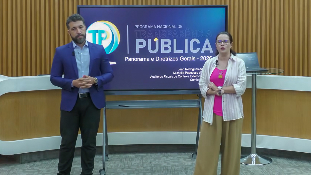 Foto horizontal dos auditores fiscais Jean Rodrigues de Souza e Michelle Padovese de Arruda, no Plenário do TCE/SC, em frente a um televisor que exibe o logo do PNTP. Eles estão em pé, com microfones. Ao fundo, há um painel de madeira.