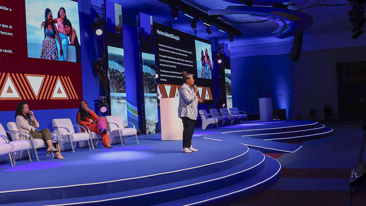 Foto da diretora Walkíria Machado Rodrigues Maciel proferindo palestra no evento. Ela é uma mulher negra, está de pé, falando ao microfone. 