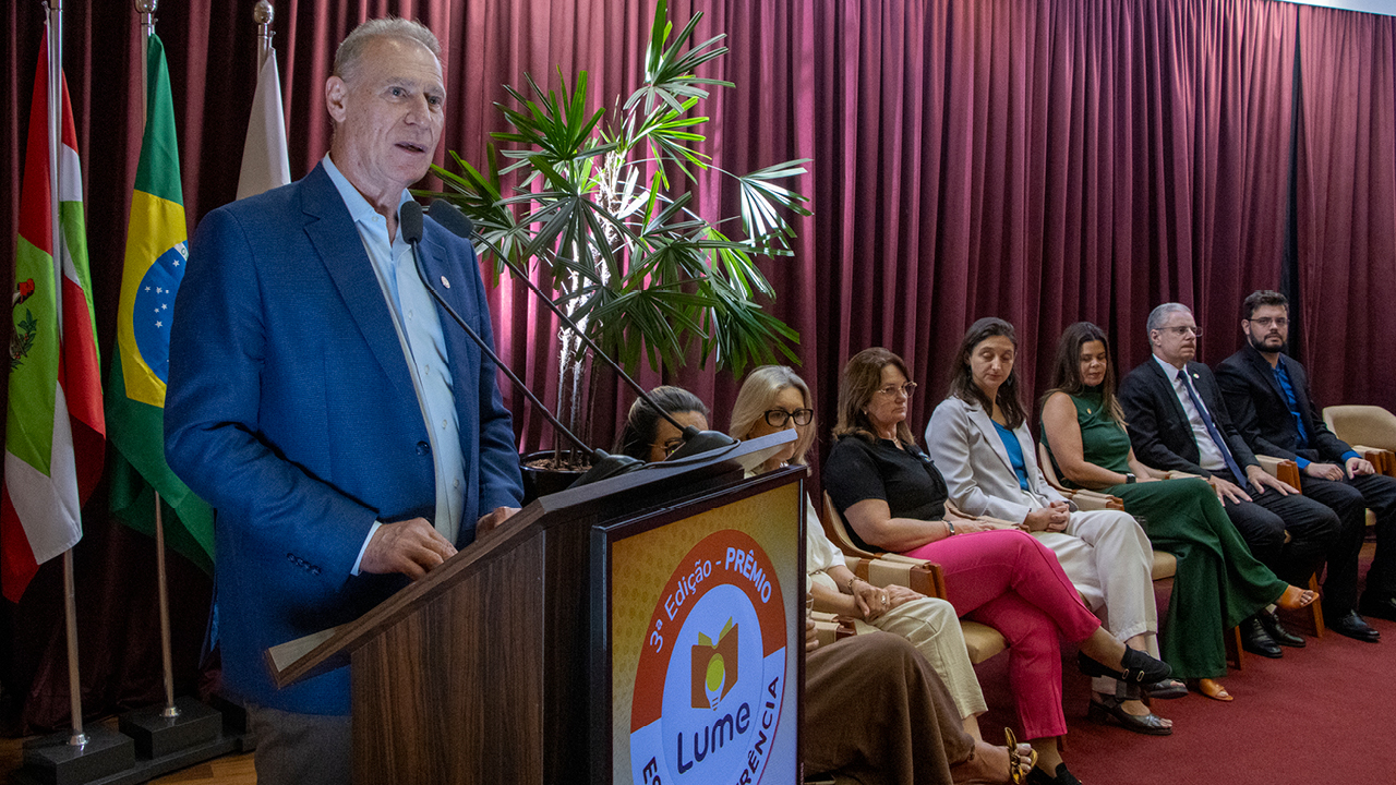 Banner horizontal com foto de várias pessoas no palco do auditório do TCE/SC. São os representantes das escolas premiadas pelo Prêmio Lume e autoridades, entre integrantes do TCE/SC, da educação do Estado e das demais instituições do acordo Lume. Alguns seguram certificados recebidos.   Banner horizontal com foto do presidente do TCE/SC, conselheiro Herneus De Nadal, discursando no púlpito durante a abertura do evento. Ele é um homem branco, de cabelos claros. Veste terno e gravata e está ao lado das demais autoridades.   Foto do conselheiro substituto Gerson Sicca falando ao púlpito no palco do auditório do TCE/SC durante a abertura do evento. Ele é um homem branco, de cabelos grisalhos. Veste terno e gravata e está ao lado da servidora do TCMRio Natália Santos.    Foto horizontal de 16 autoridades no palco do auditório durante o evento. Entre os presentes, conselheiros do TCE/SC, representantes da educação do Estado e de demais instituições do acordo Lume.  