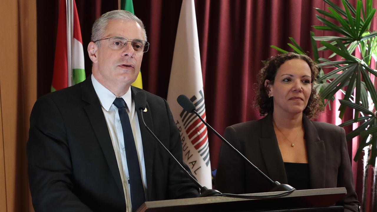 Foto do conselheiro substituto Gerson Sicca falando ao púlpito no palco do auditório do TCE/SC durante a abertura do evento. Ele é um homem branco, de cabelos grisalhos. Veste terno e gravata e está ao lado da servidora do TCMRio Natália Santos. 
