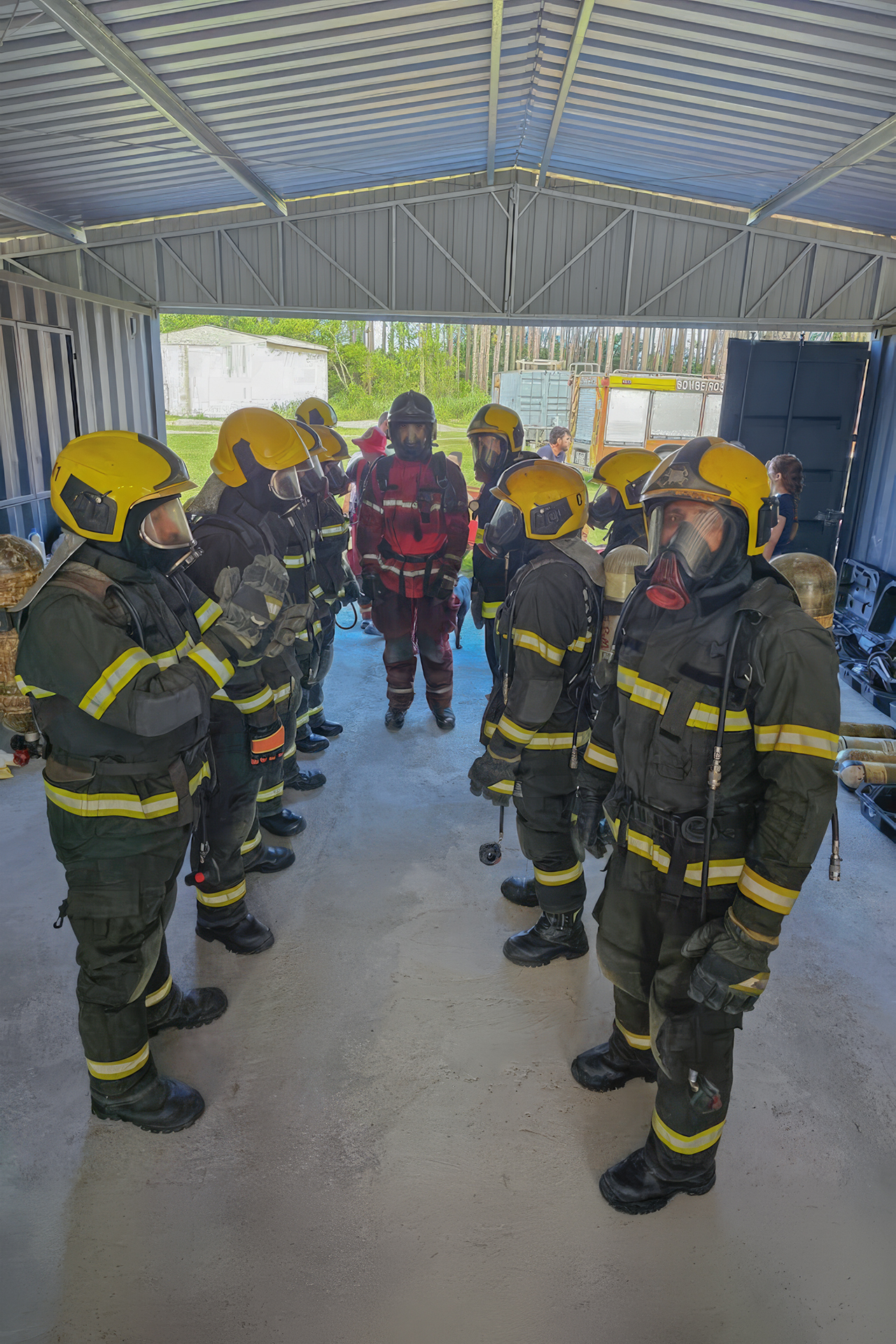 Imagem em ambiente coberto, semelhante a um galpão, mostrando a 2ª turma da brigada de incêndio do TCE/SC durante treinamento. Ao centro, um instrutor com uniforme vermelho e equipamentos de proteção orienta o grupo. Ao redor, brigadistas formam duas fileiras frente a frente, vestindo trajes completos de combate a incêndio, com capacetes amarelos, máscaras respiratórias e roupas com faixas refletivas.