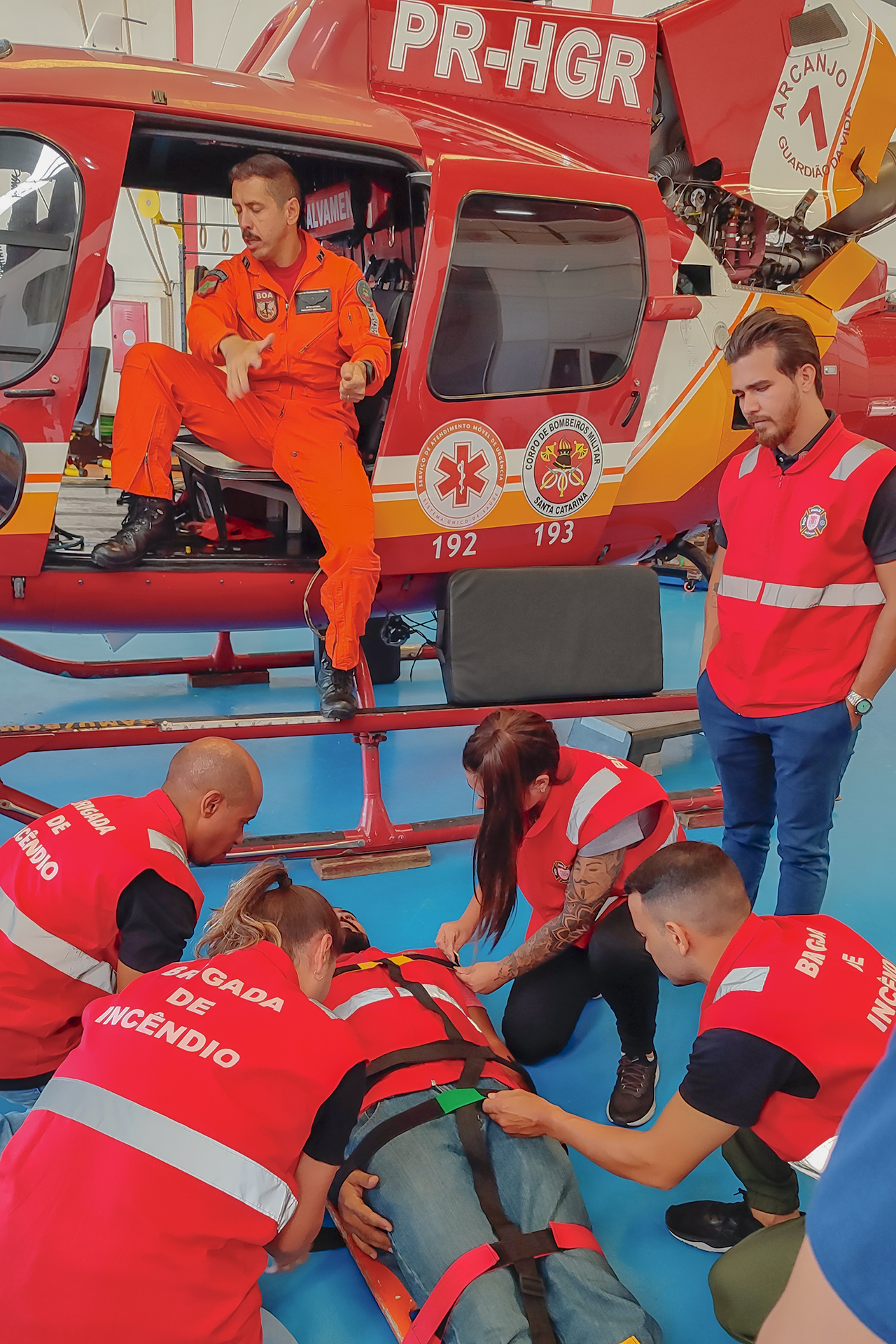 Imagem em ambiente de treinamento da 2ª turma da brigada de incêndio do TCE/SC. Em primeiro plano, brigadistas utilizam equipamentos de imobilização para atender uma pessoa deitada, simulando resgate de vítima. Ao redor, integrantes da equipe, identificados com coletes vermelhos da brigada de incêndio, prestam apoio à ação. Ao fundo, há um helicóptero de resgate com a porta aberta, e um profissional uniformizado em macacão laranja observa a atividade. 