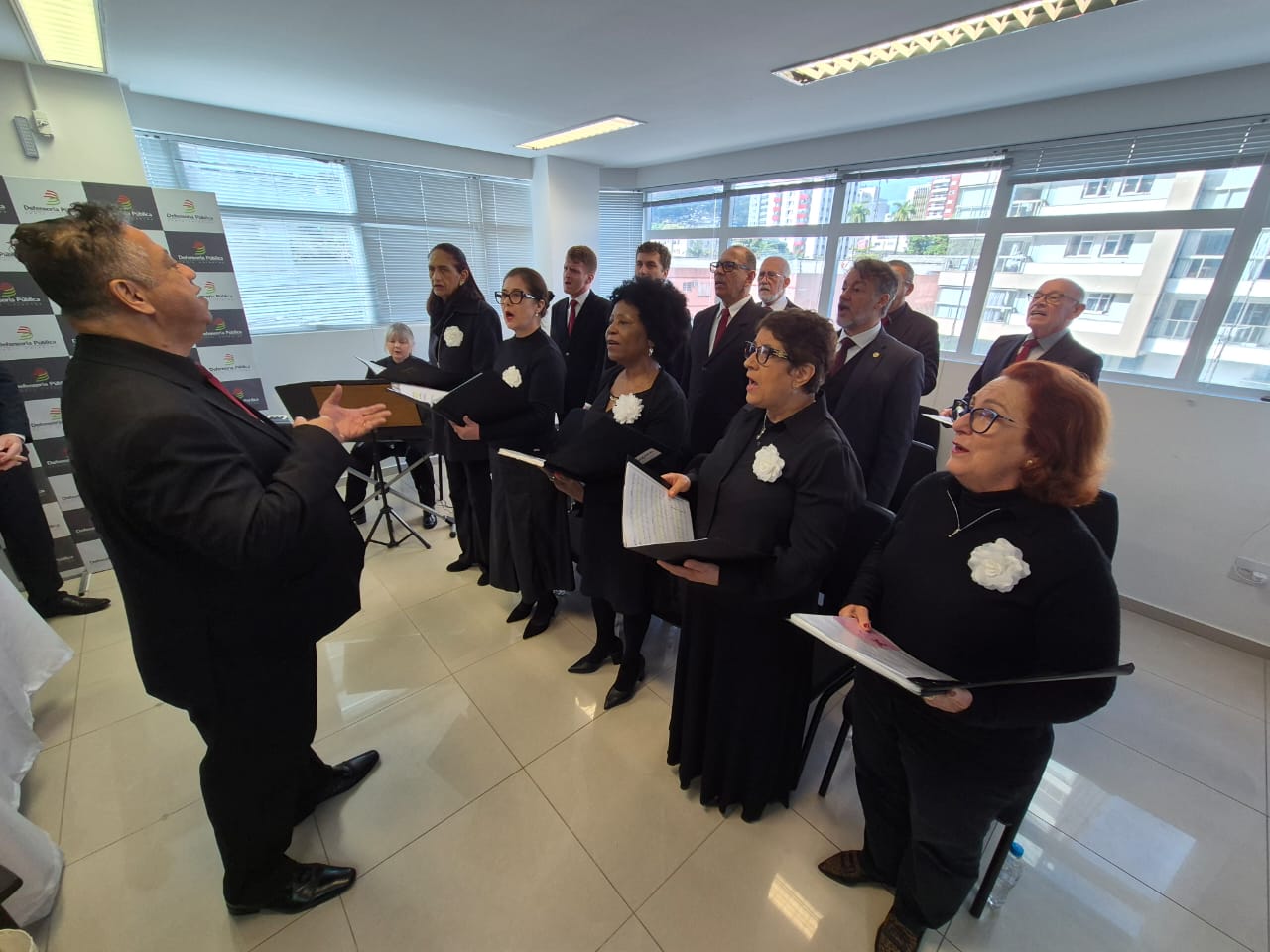 Foto do Coral Hélio Teixeira da Rosa durante apresentação no evento. O maestro está à frente, conduzindo os demais. Há várias pessoas agrupadas, cantando. Todos vestem roupa preta. 