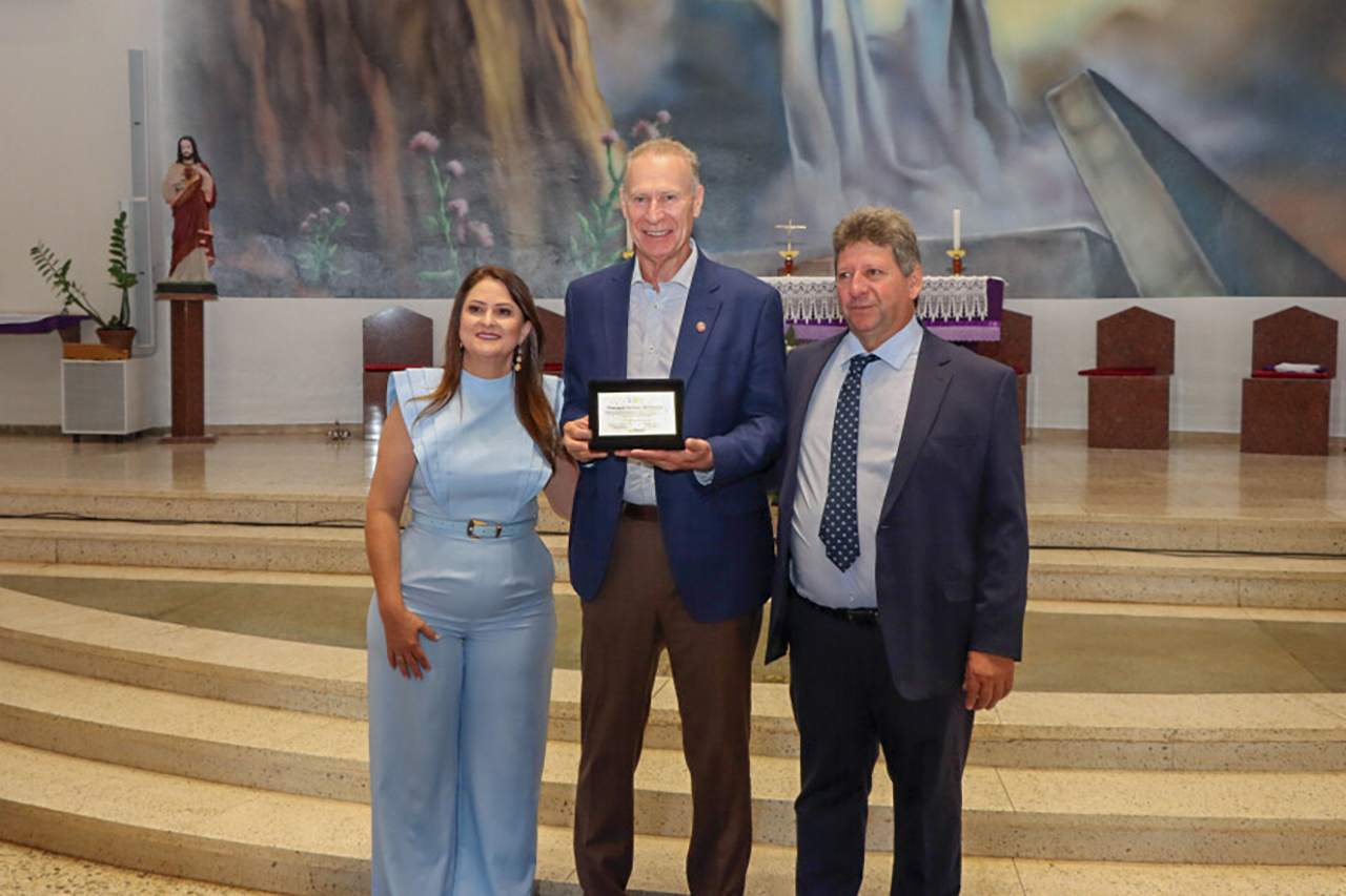 Imagem horizontal com foto em ambiente interno com escadaria e painel artístico ao fundo, mostrando três pessoas posando para registro oficial; ao centro, o presidente Herneus De Nadal segura uma placa de homenagem. Ao seu lado, a prefeita e o vice-prefeito de Palmitos, Giovana Giacomolli e Itamar Fiorese. 