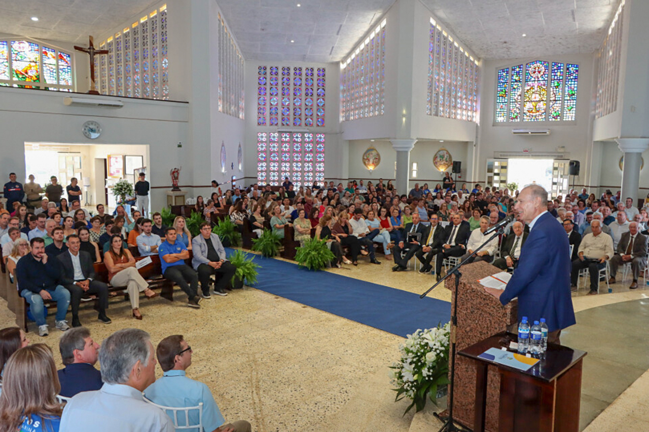 Foto horizontal em ambiente interno amplo, com vitrais coloridos e altar ao fundo, mostrando o presidente Herneus De Nadal discursando em púlpito com microfone para um grande público acomodado em bancos e cadeiras. 