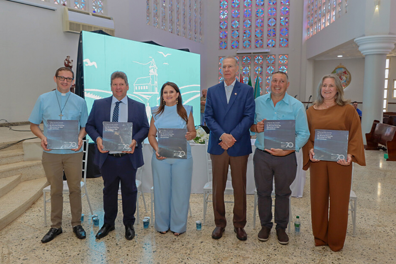 Imagem horizontal de seis pessoas em ambiente interno, posicionadas lado a lado, voltadas para a câmera. Cinco delas seguram o livro comemorativo aos 70 anos do TCE/SC. Ao centro, o presidente Herneus De Nadal está em pé, usando blazer azul e camisa clara. Entre os demais presentes, a prefeita e o vice-prefeito de Palmitos, Giovana Giacomolli e Itamar Fiorese. 