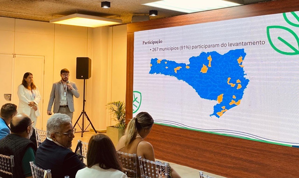 Foto dos servidores Elisete e Mateus falando ao público durante o evento. Eles estão em pé, ao fundo, próximo ao telão que projeta sua apresentação. À frente, há pessoas sentadas em cadeiras, assistindo à fala.