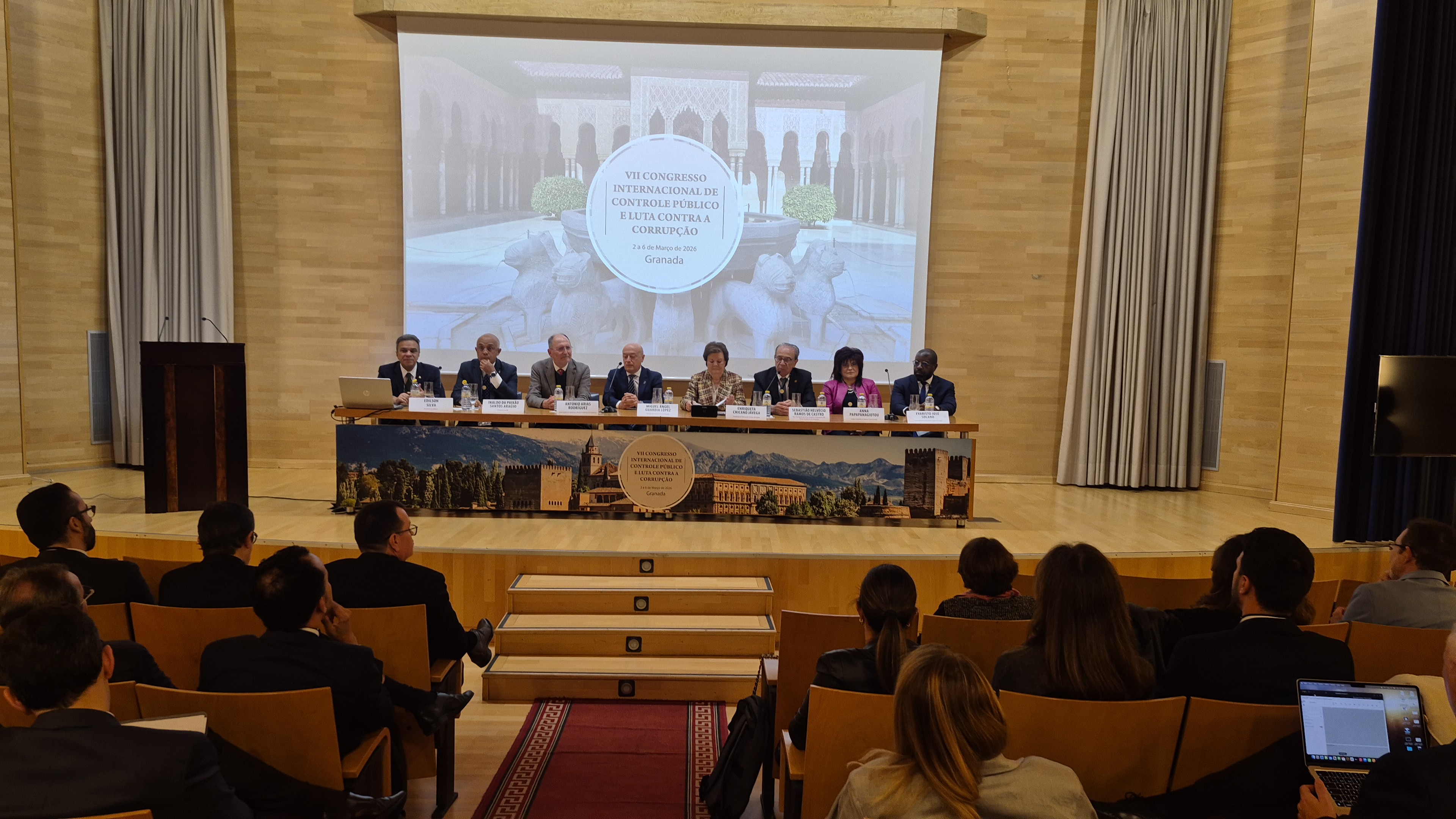 Foto horizontal da bancada de autoridades durante a abertura do evento, no palco. Os participantes estão lado a lado, identificados por placas nominais. Há oito pessoas. Entre elas, o presidente da Atricon, conselheiro Edilson Silva. Ao fundo, telão que exibe o nome do congresso. À frente do palco, de costas, a plateia. 