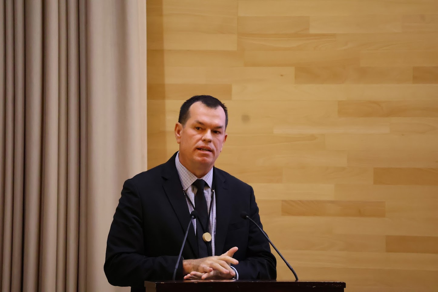 Foto do conselheiro Adircélio de Moraes Ferreira Júnior falando no púlpito durante o evento. Ele é um homem branco, de cabelos escuros. Veste terno e gravata e usa uma medalha dourada no pescoço. Ao fundo, há cortinas. 