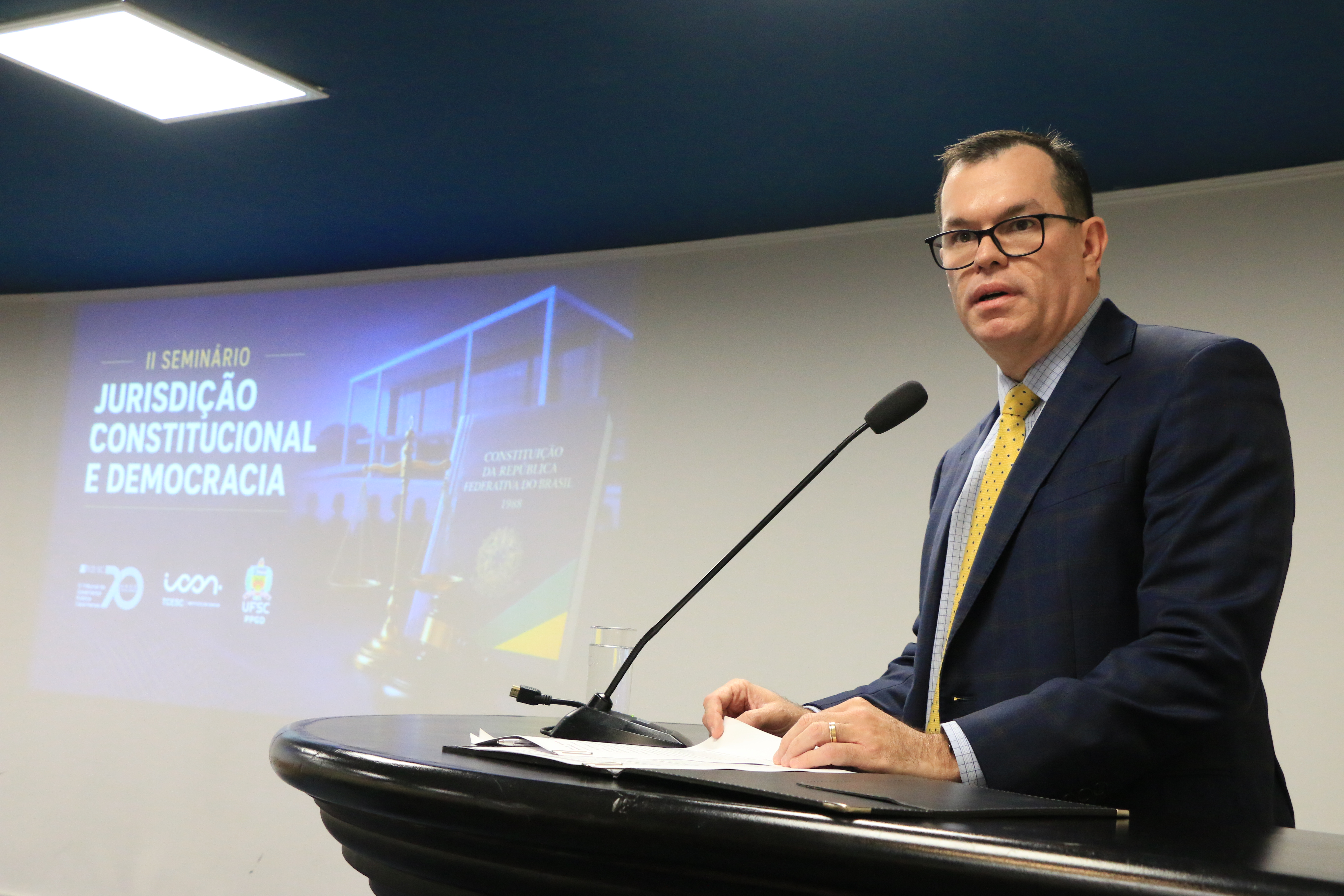 Foto horizontal do conselheiro Adircélio Ferreira Júnior falando no púlpito do auditório. Ele é um homem branco, de cabelos escuros. Veste terno e gravata e usa óculos de grau. Ao fundo, no telão, projeção do logo do evento.