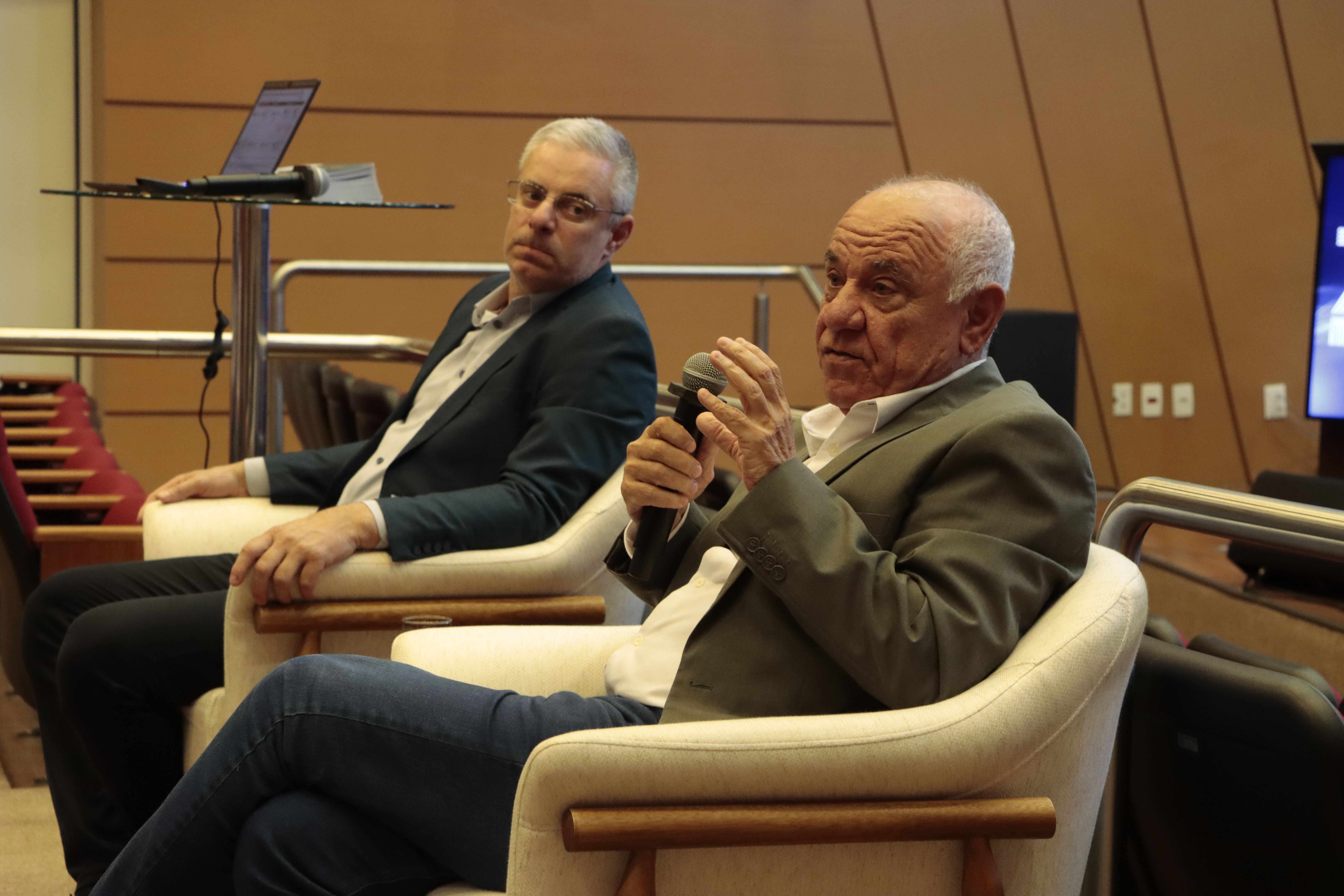 Foto horizontal do conselheiro Luiz Eduardo Cherem falando ao microfone no auditório do TCE/SC. Ele está sentado em poltrona, ao lado do conselheiro Sicca.