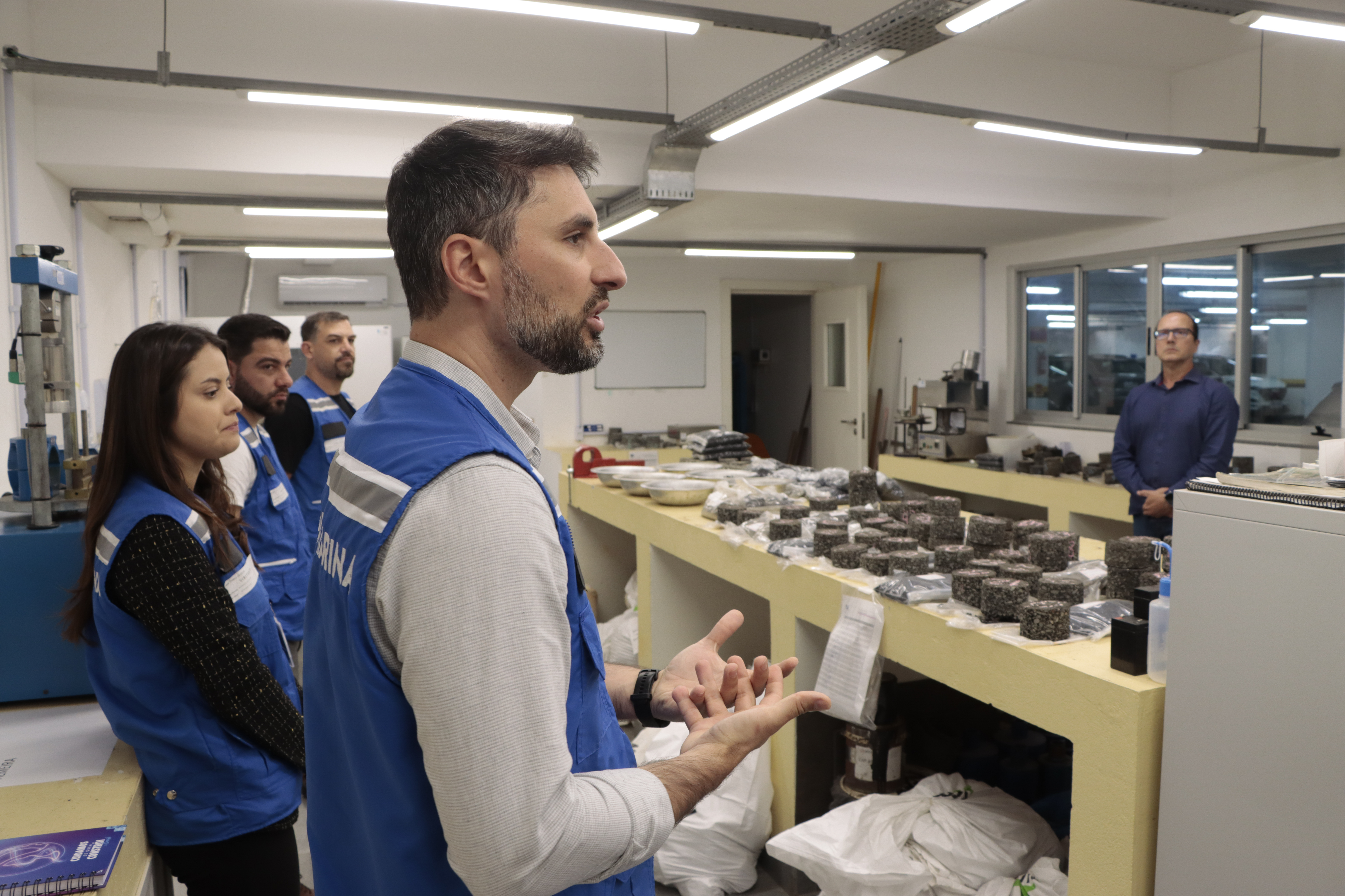 Foto horizontal que mostra, em primeiro plano, o gerente do Setor de Infraestrutura do Cincatarina, Maurício Perazzoli, falando aos demais durante visita ao laboratório do TCE. Ele está ao lado de outros colegas. À sua frente, uma mesa com vários materiais e equipamentos de obras rodoviárias.