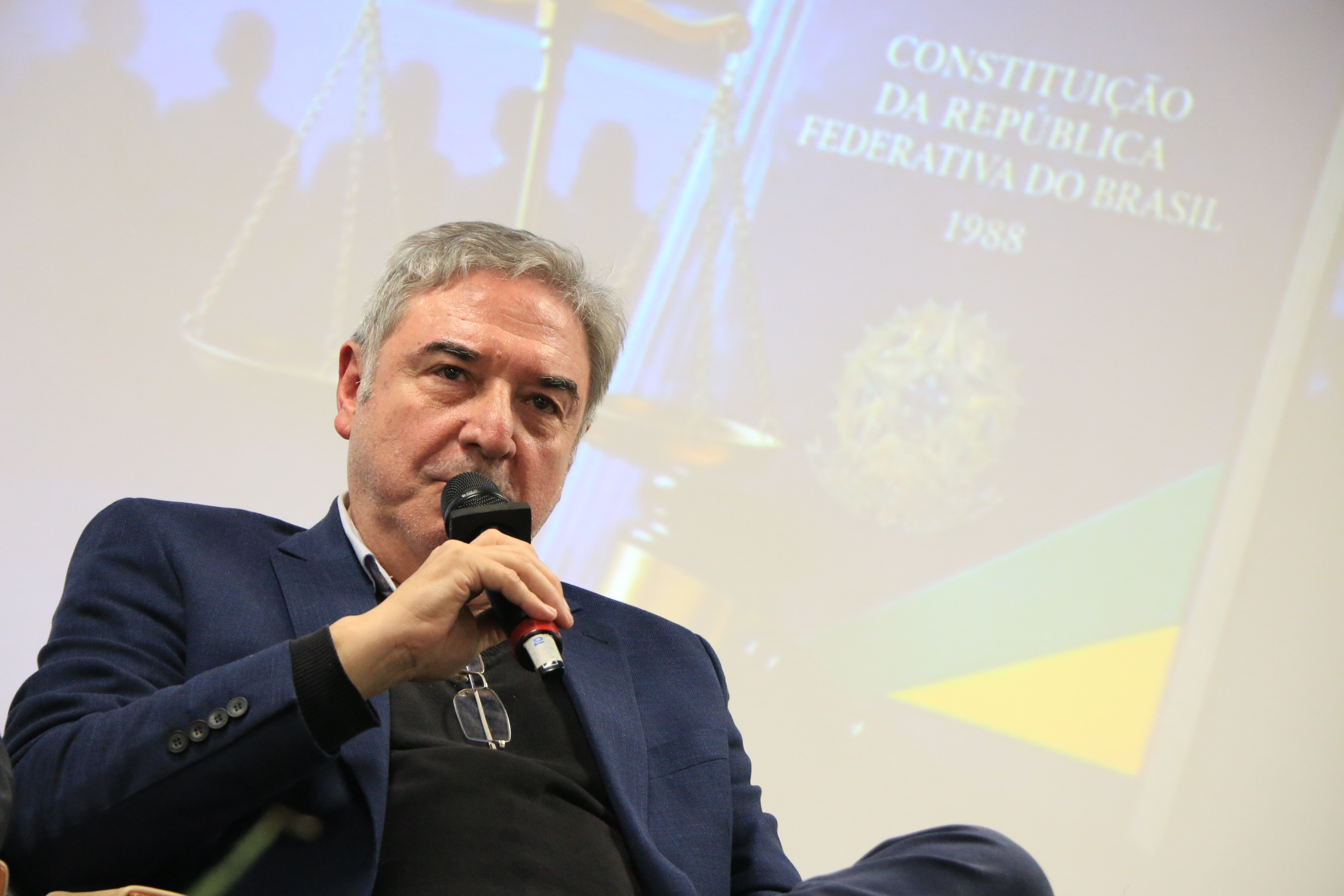 Foto horizontal do professor Luiz Henrique Cademartori falando ao microfone durante evento. É um homem branco, de cabelos grisalhos. Ao fundo, no telão, aparece exemplar da Constituição.