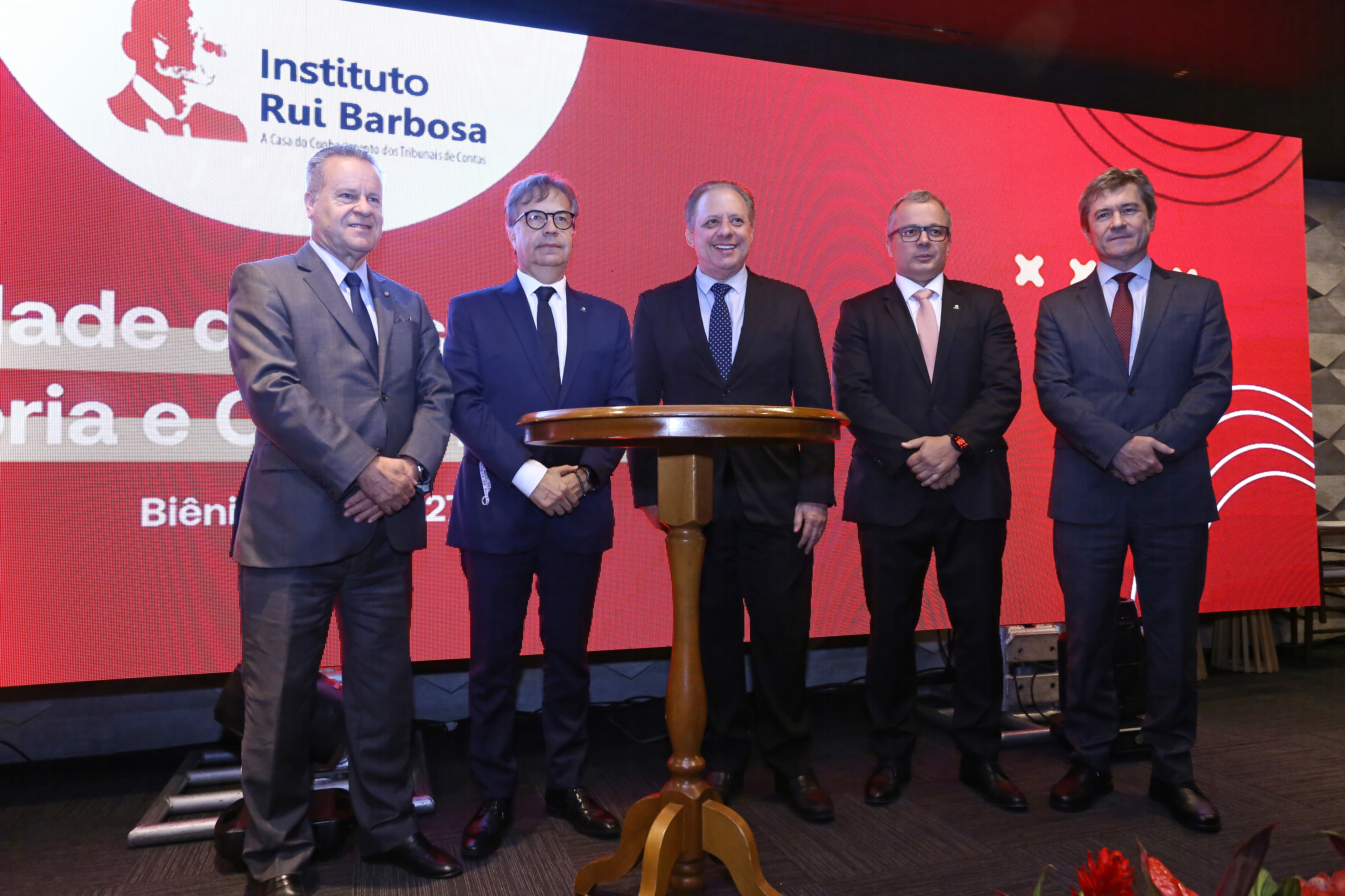Banner horizontal com foto da diretoria do IRB juntamente com o conselheiro José Nei Ascari. Há cinco homens, lado a lado, durante o evento. Estão todos em pé, no palco, vestindo terno e gravata. Ao fundo, no telão, logo do IRB.  