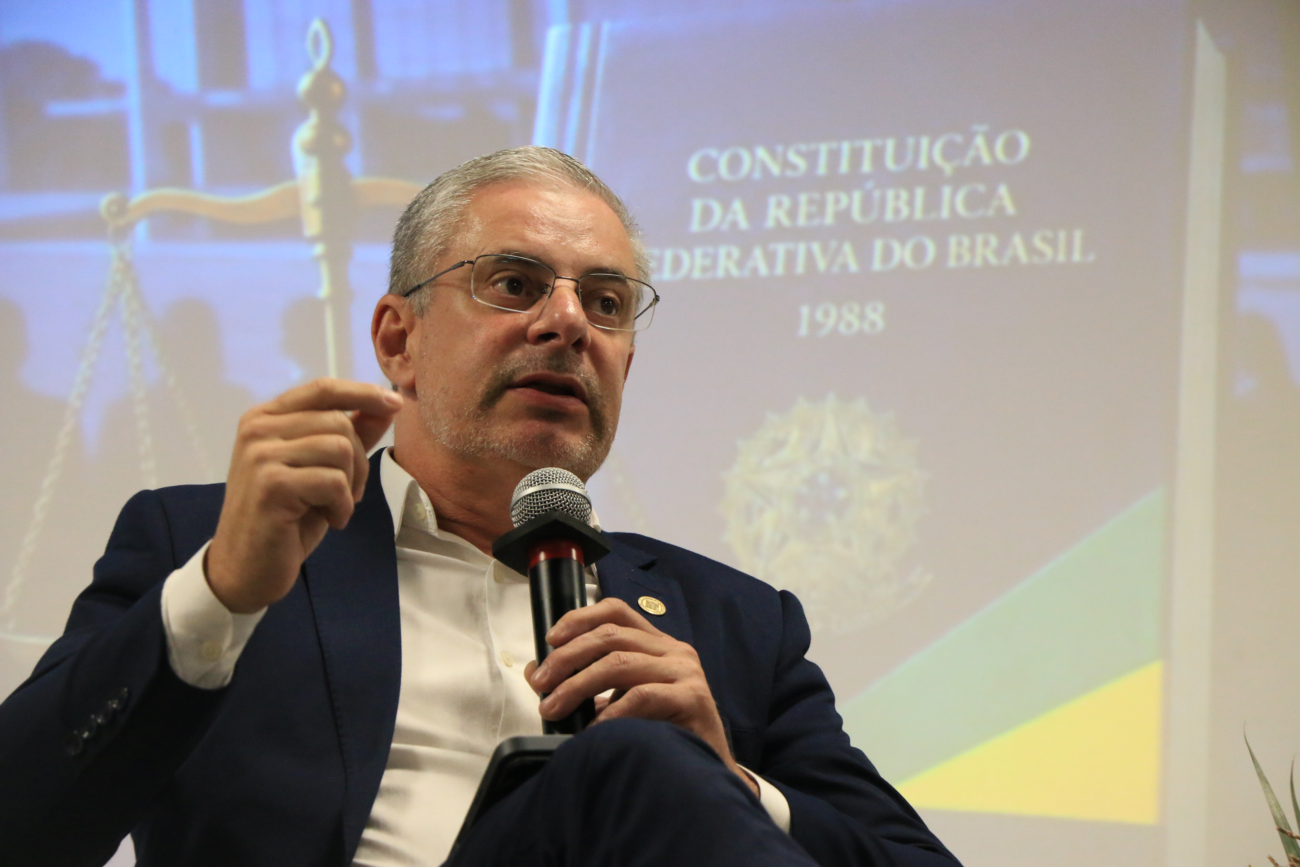 Foto horizontal do conselheiro substituto Gerson Sicca falando ao microfone durante evento. Ele é um homem branco, de cabelos claros. Veste terno e gravata e usa óculos de grau. Ao fundo, no telão, aparece imagem de exemplar da Constituição.