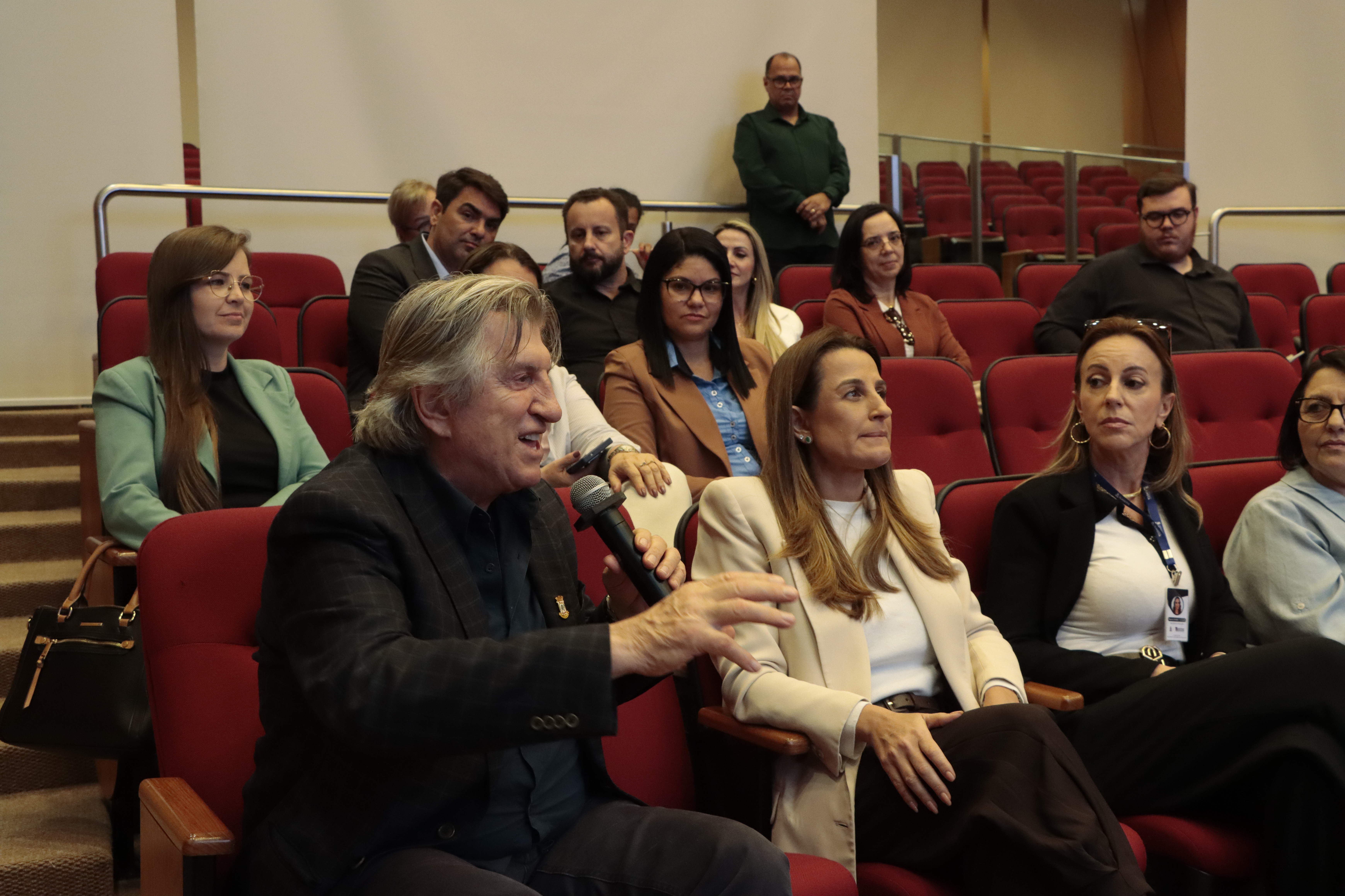 Foto horizontal de um membro da plateia fazendo uma pergunta ao microfone. Ele está sentado em poltrona no auditório, ao lado de outras pessoas. Entre os presentes, os prefeitos Leonel Pavan e Juliana Pavan.
